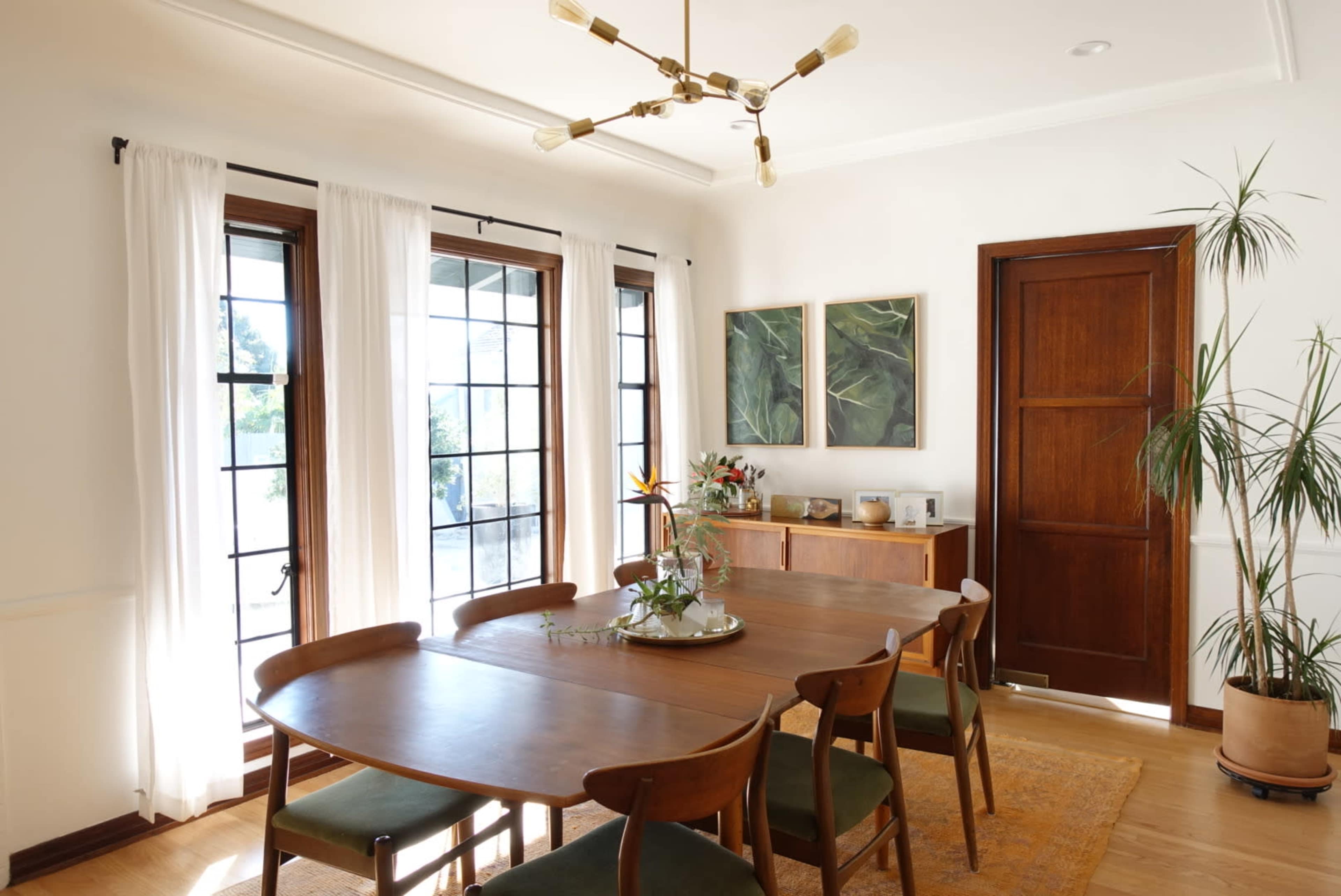 A dining room features a wooden table surrounded by chairs, large windows with sheer curtains, and minimalist decor including plants and artwork.