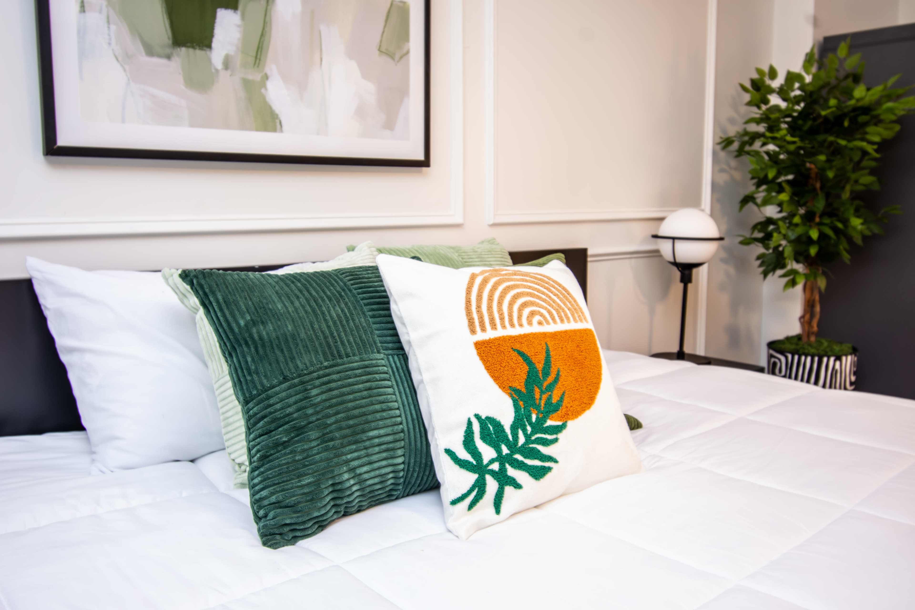 A bed with decorative pillows and a potted plant is arranged in a room featuring light-colored walls and framed artwork.