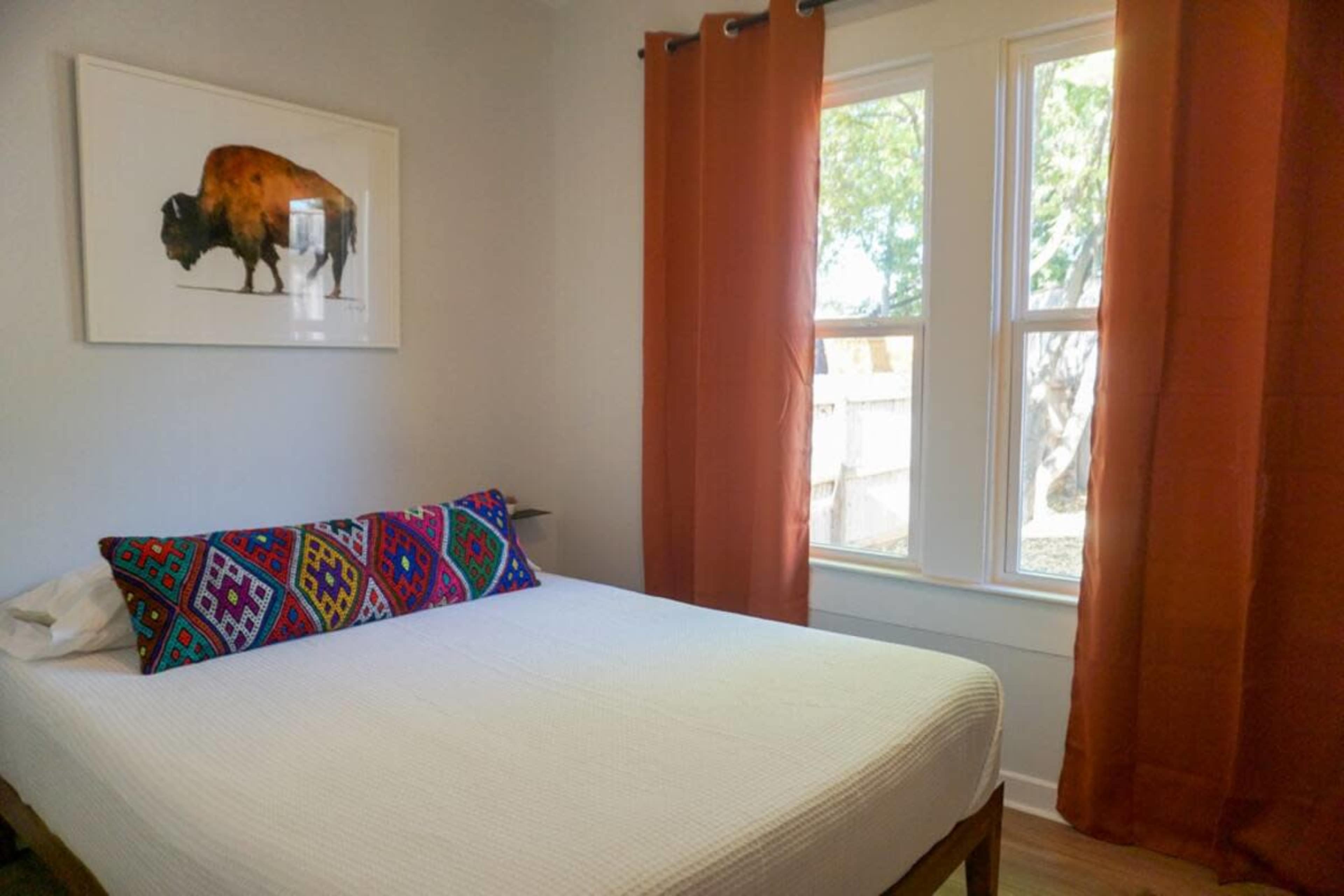 A bedroom with a bed featuring a colorful patterned pillow and orange curtains framing two windows.