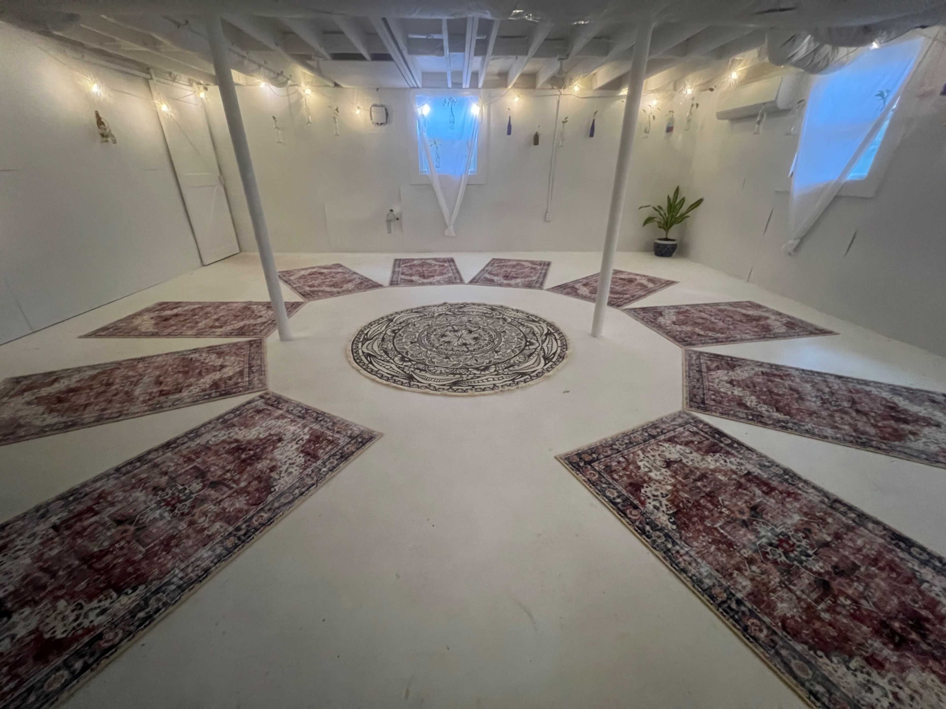A wide, empty basement room features a central circular rug surrounded by several smaller rugs arranged in a circular pattern, with soft lighting from multiple sources.