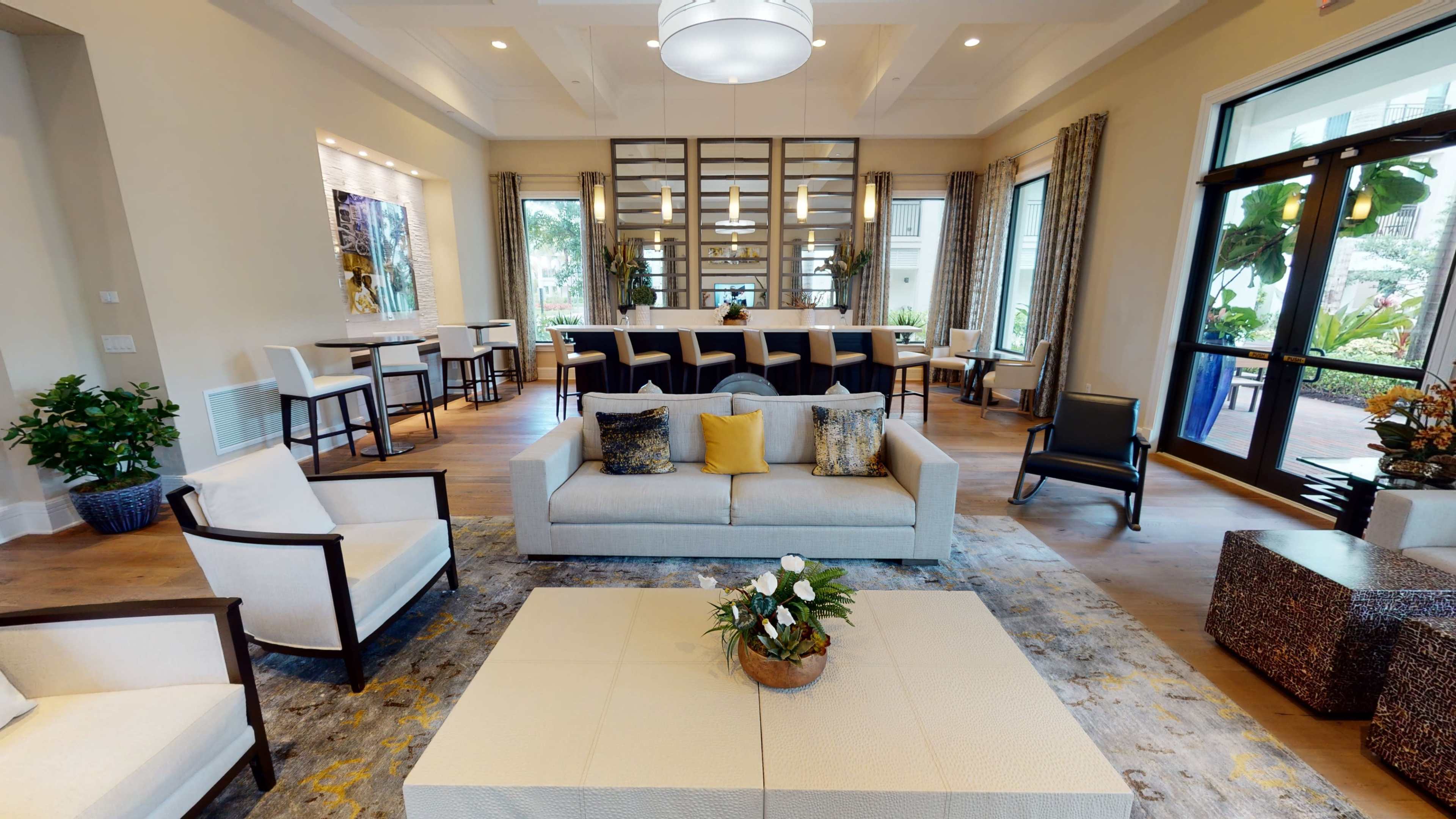 The image shows a modern lounge area with a light-colored sofa, several seating options, a coffee table, and large windows allowing natural light.