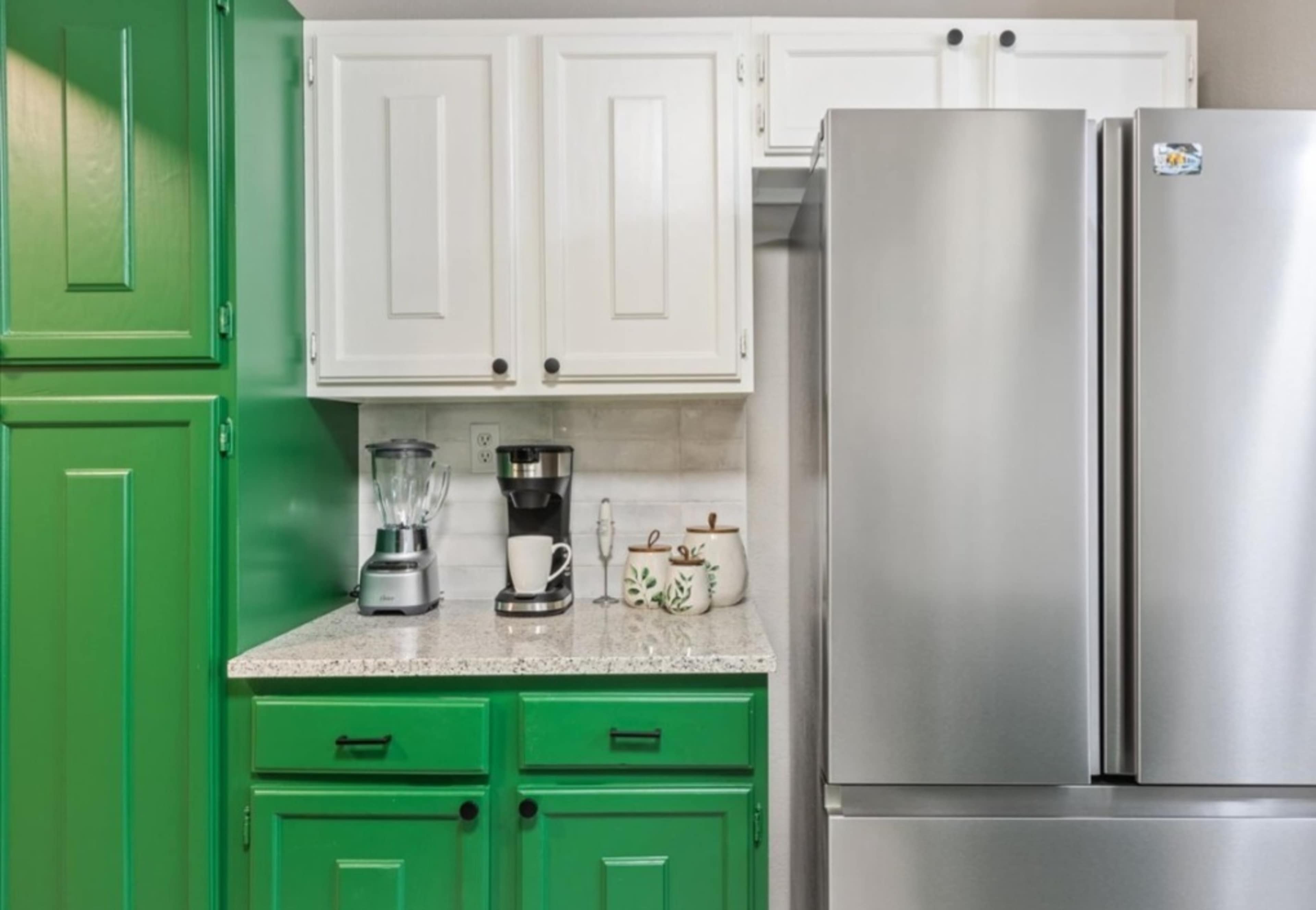 A kitchen corner with green cabinets, white upper cabinets, a silver refrigerator, and countertop appliances including a blender and coffee maker.