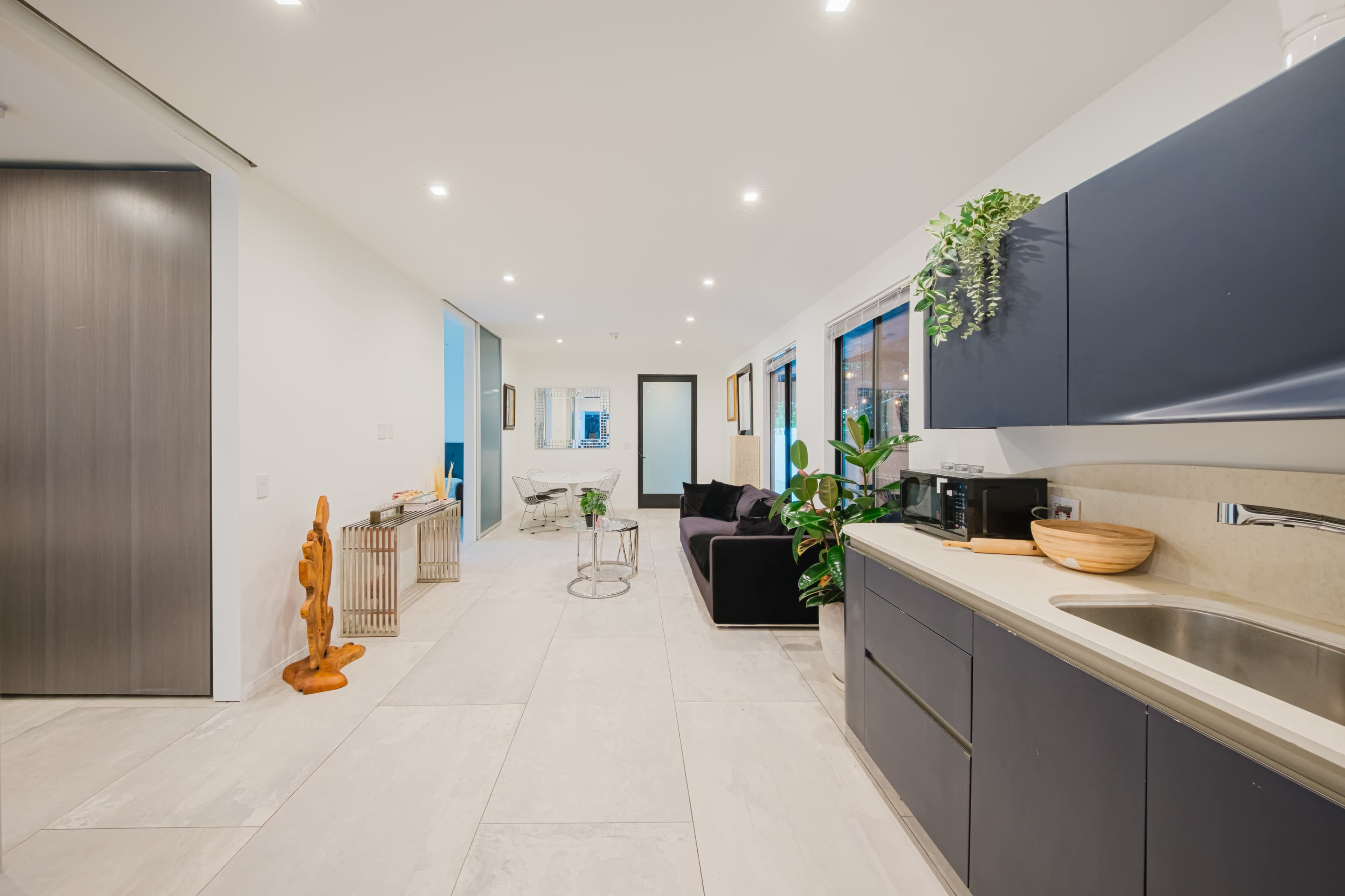 The image shows a modern, open-concept living space featuring a kitchen with dark cabinetry, a dining area, and a sitting area with a black couch.