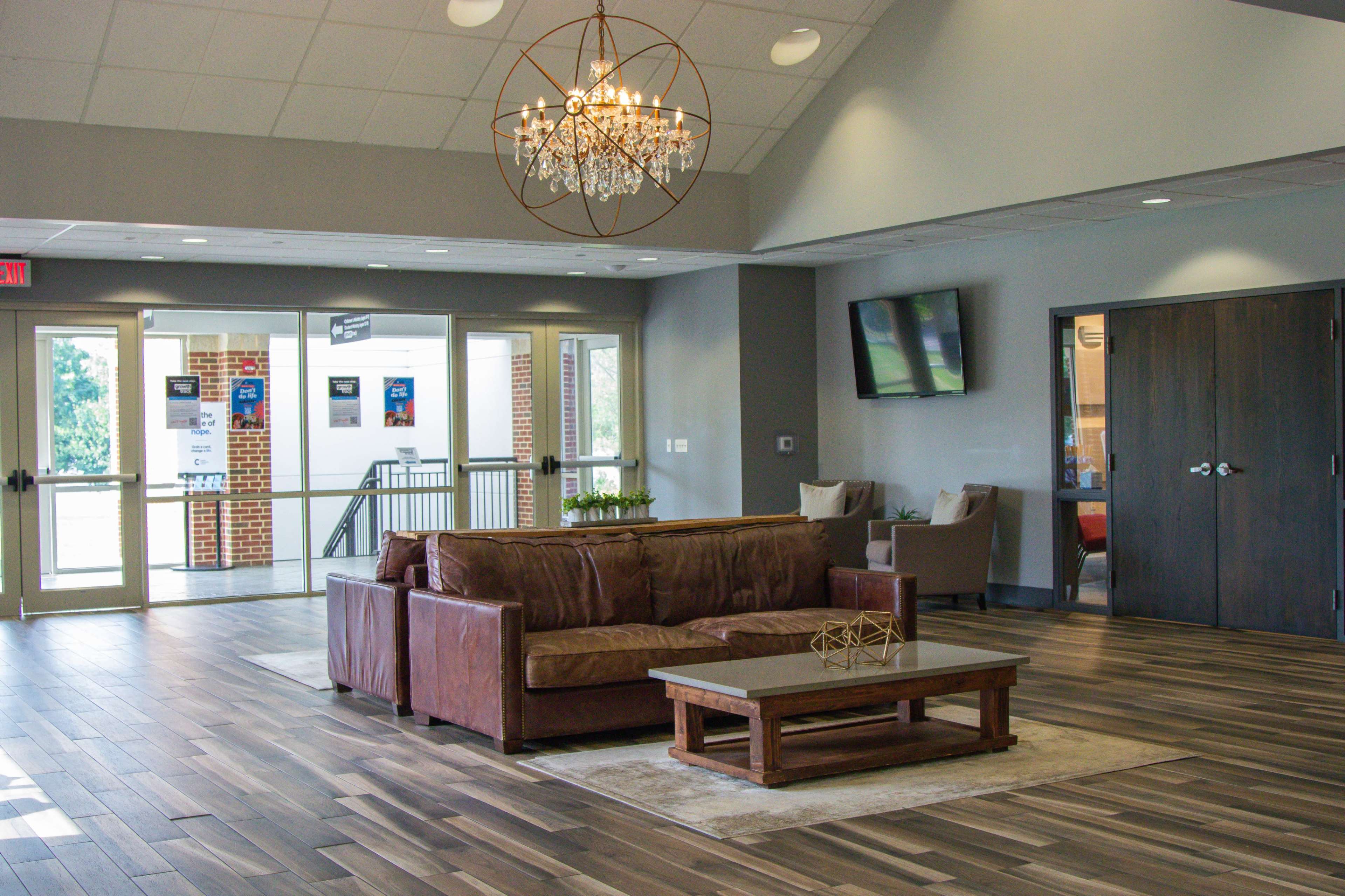 A modern lobby area featuring a brown leather sofa, a wooden coffee table, and a chandelier, with large windows and a glass door leading outside.