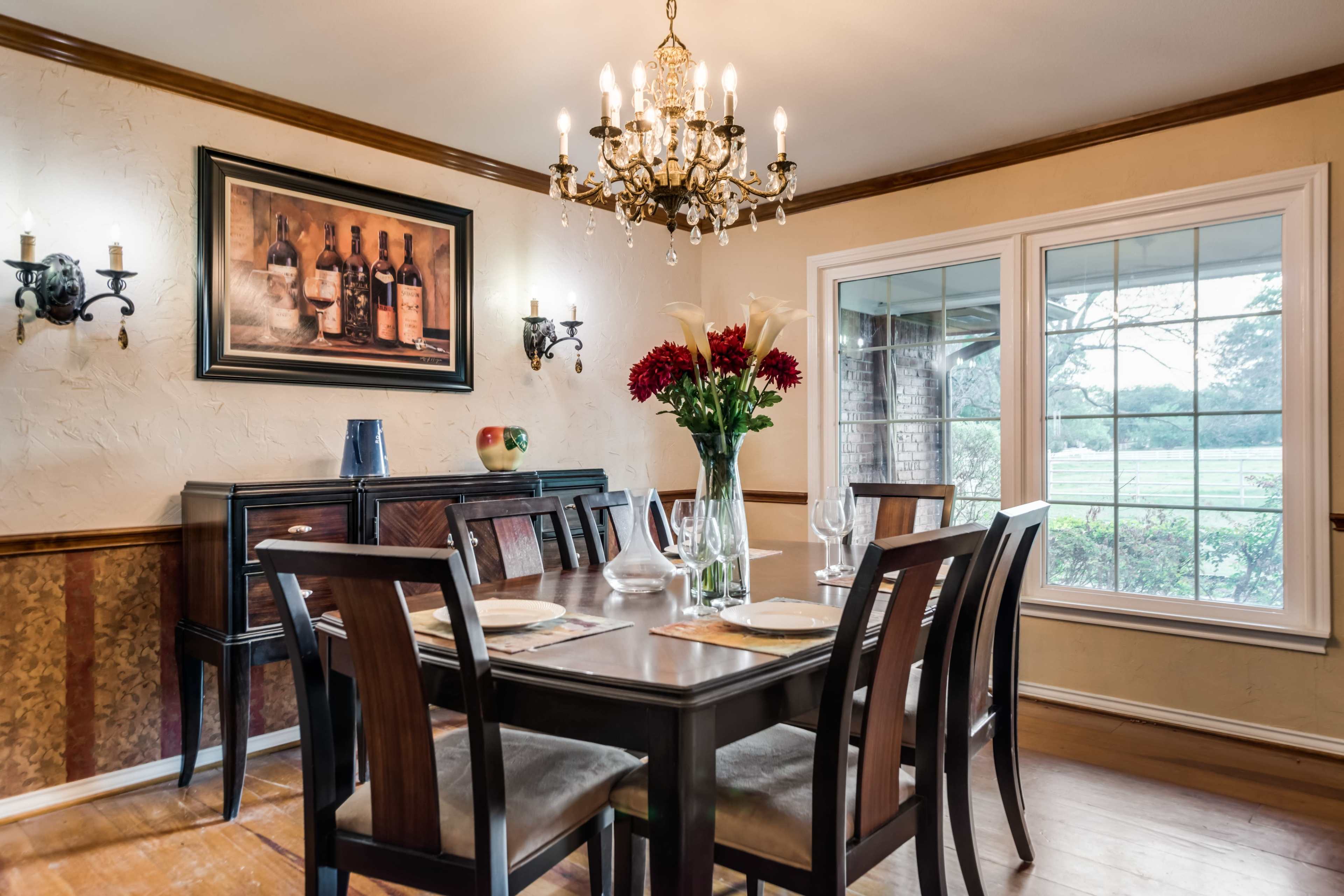 A dining room features a wooden table surrounded by black chairs, with a chandelier above and a large window overlooking the outdoors.