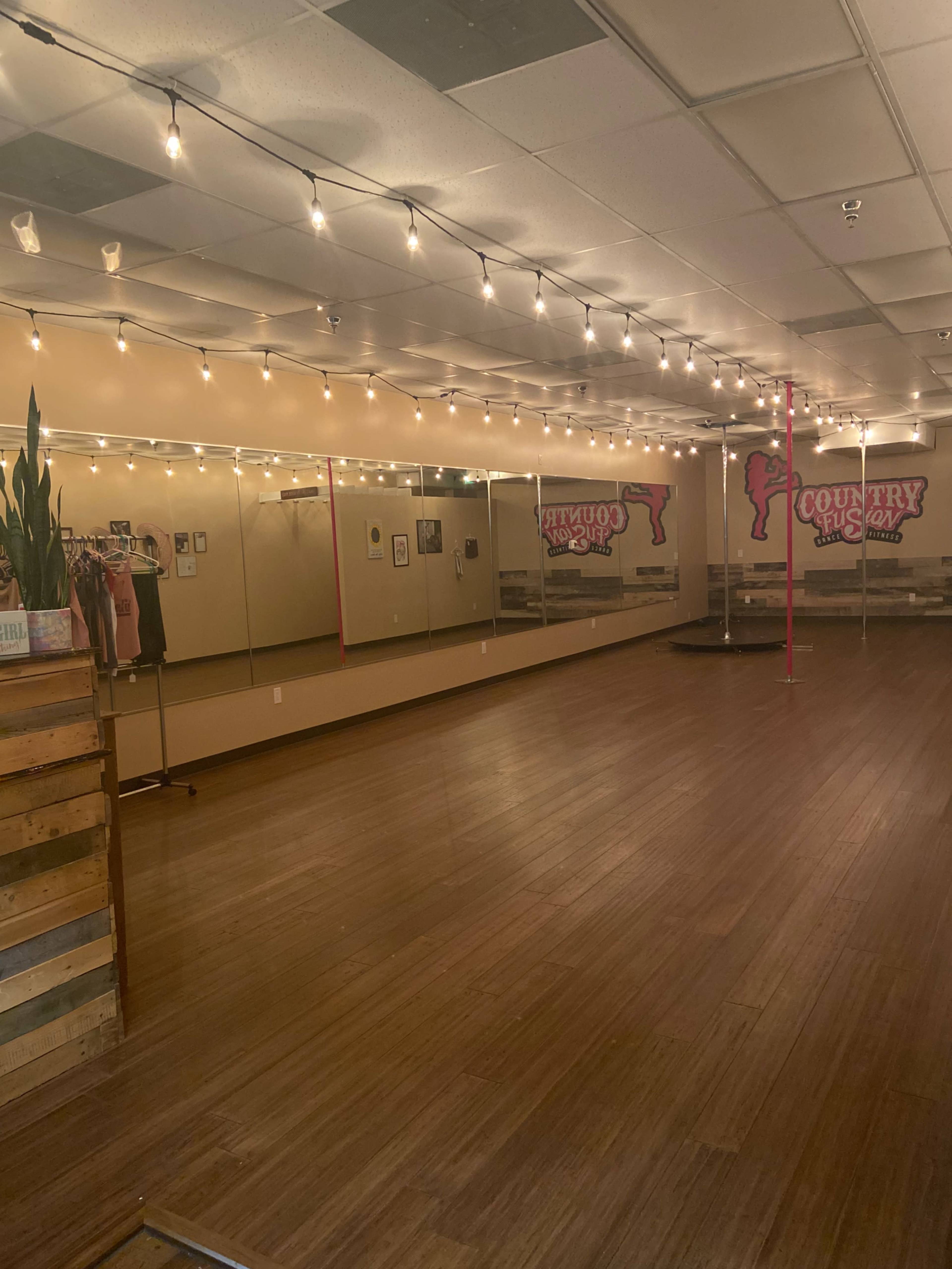 An empty dance studio with wooden flooring, mirrors on one wall, and string lights overhead.