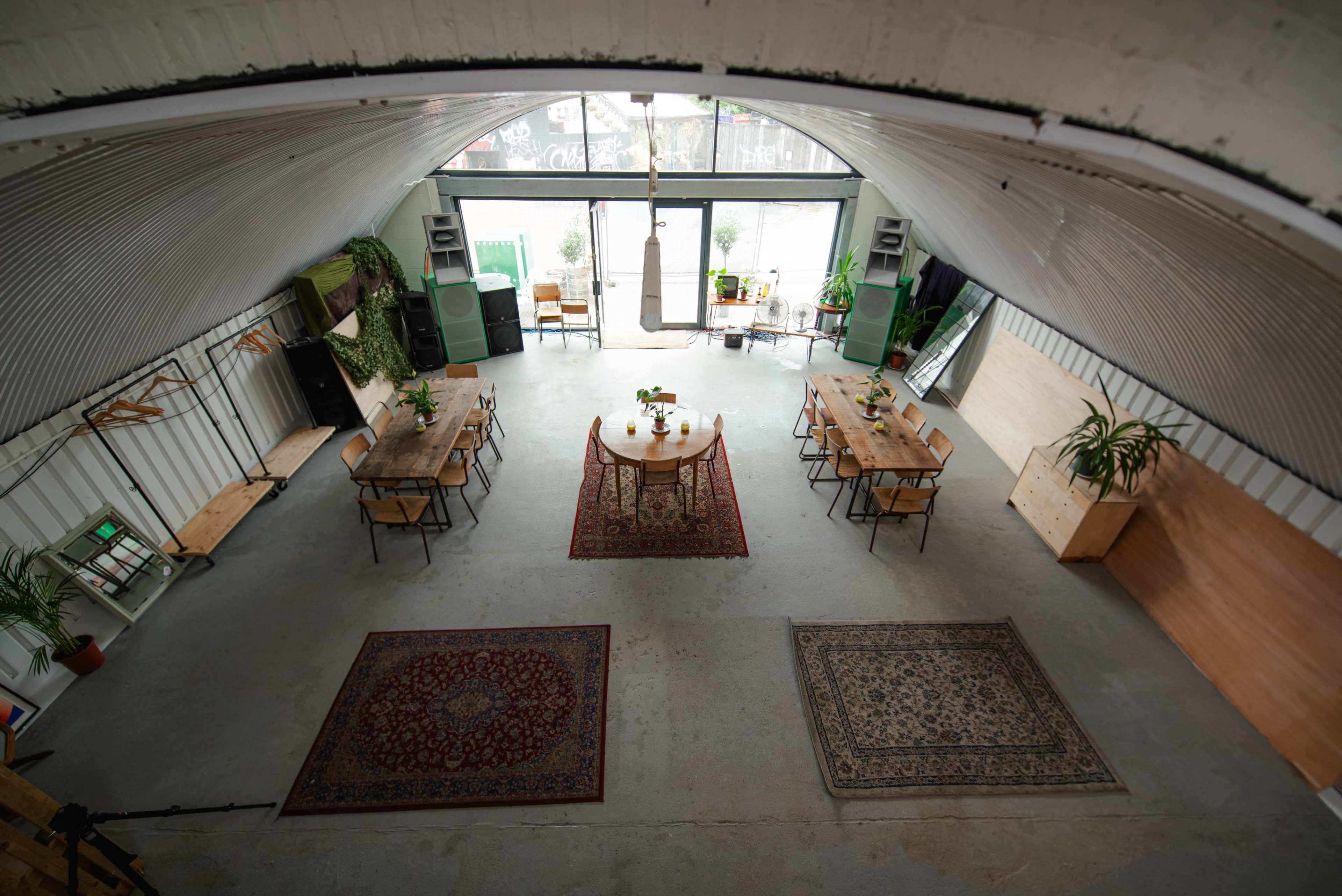 The image shows a spacious indoor area with several wooden tables, chairs, and decorative rugs, along with plants, a large window letting in natural light, and an industrial-style ceiling.