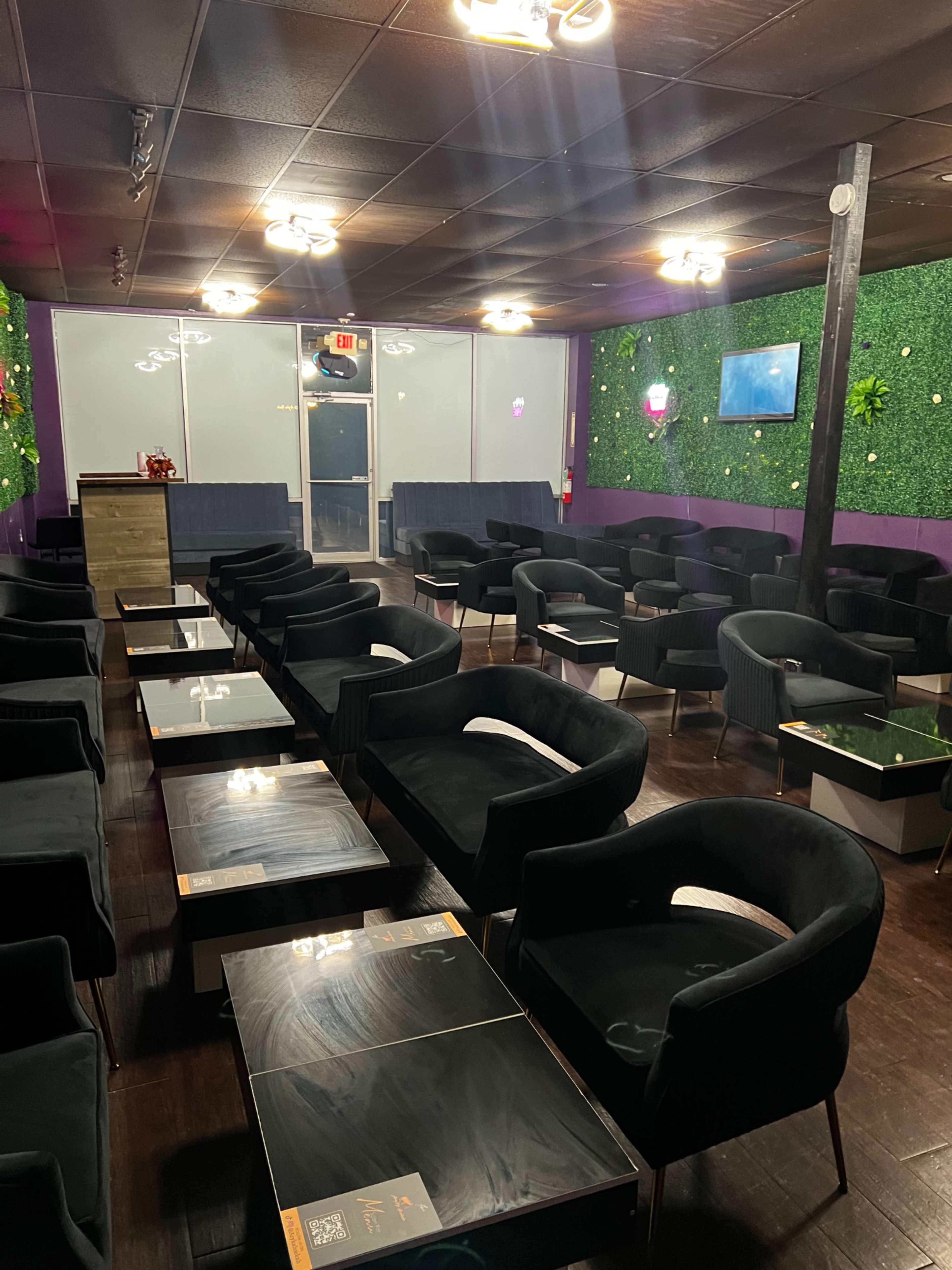 A lounge area with several black chairs and tables arranged in rows, surrounded by green wall decor and dim lighting.