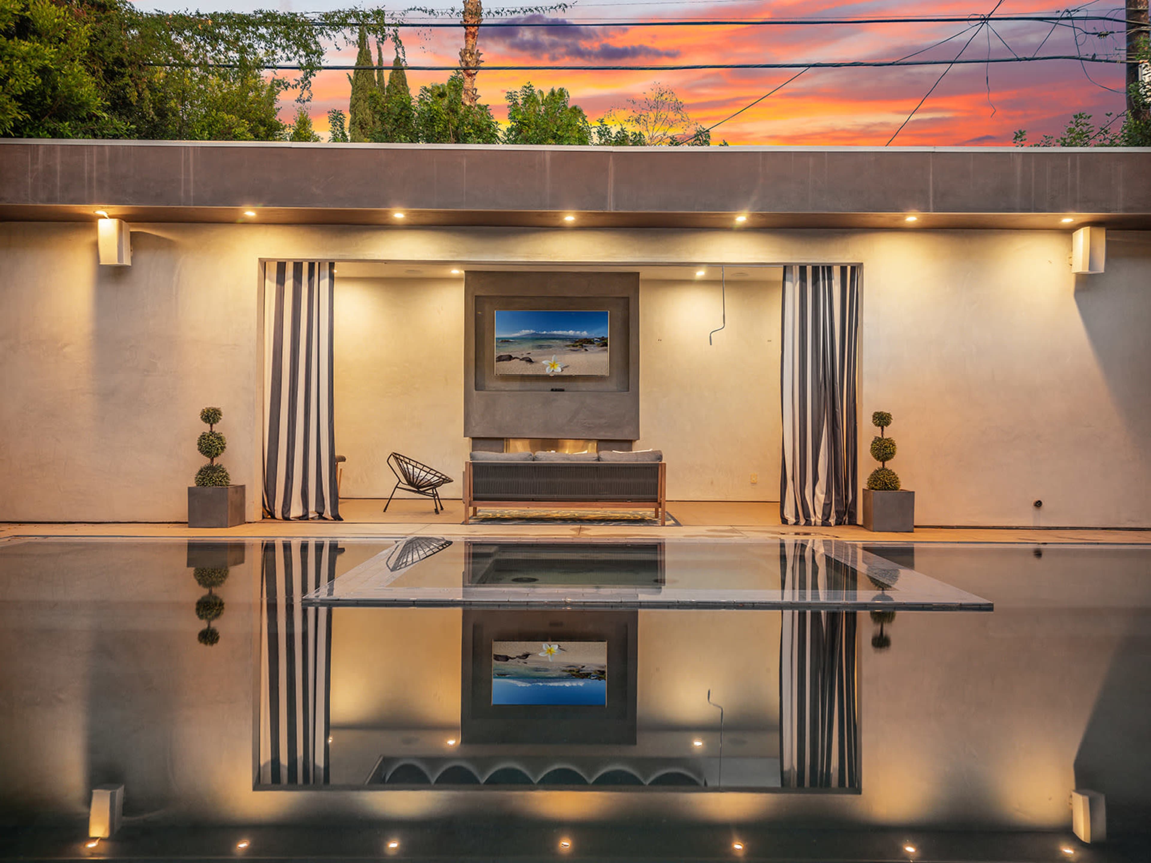 A modern outdoor space features a pool reflecting a minimalist lounge area with a large screen and potted plants, set against a colorful sunset sky.