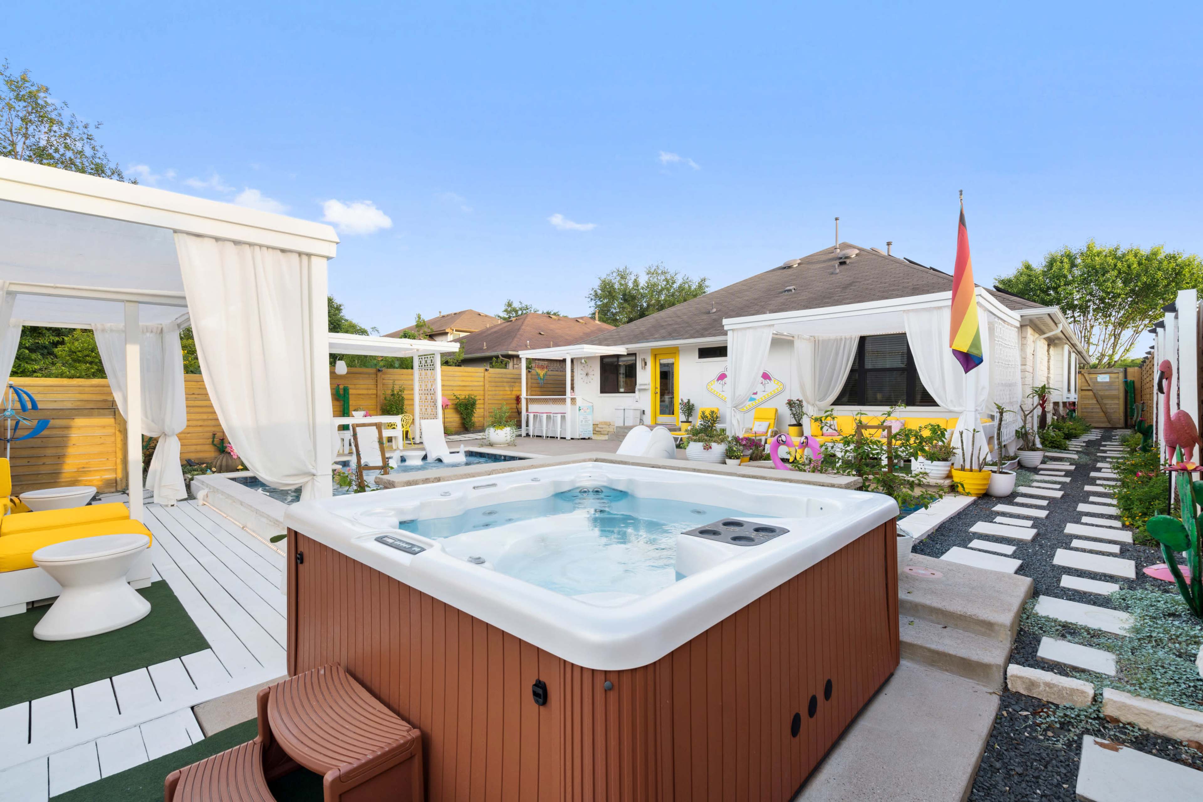 The image shows a backyard featuring a hot tub, seating areas, and a well-maintained garden with colorful flowers and pathways leading to a house.