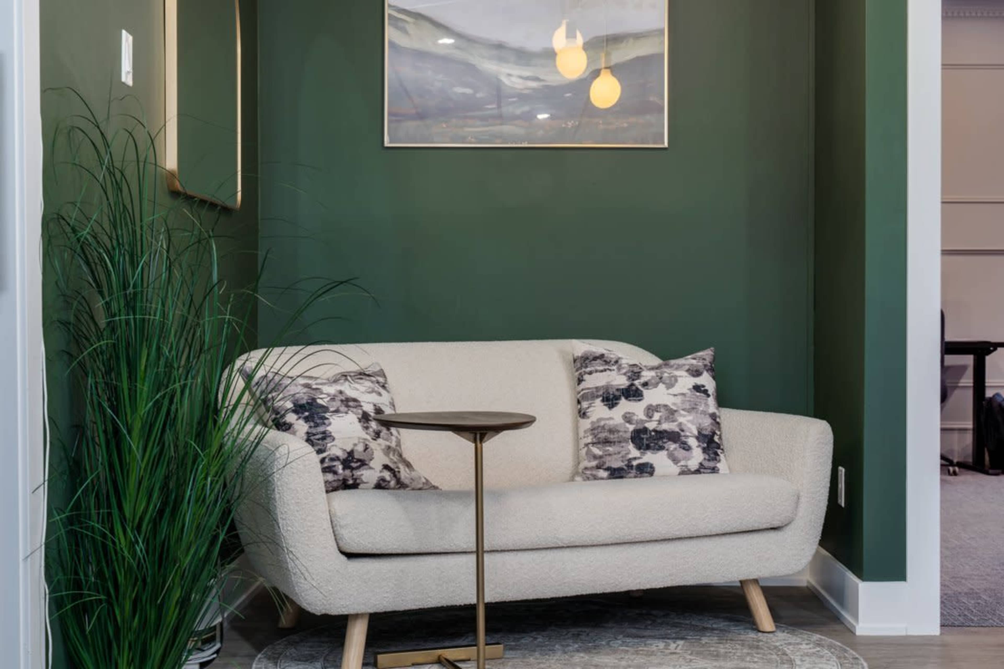 A small couch with decorative pillows sits next to a round side table in a room with dark green walls and a piece of art on the wall.