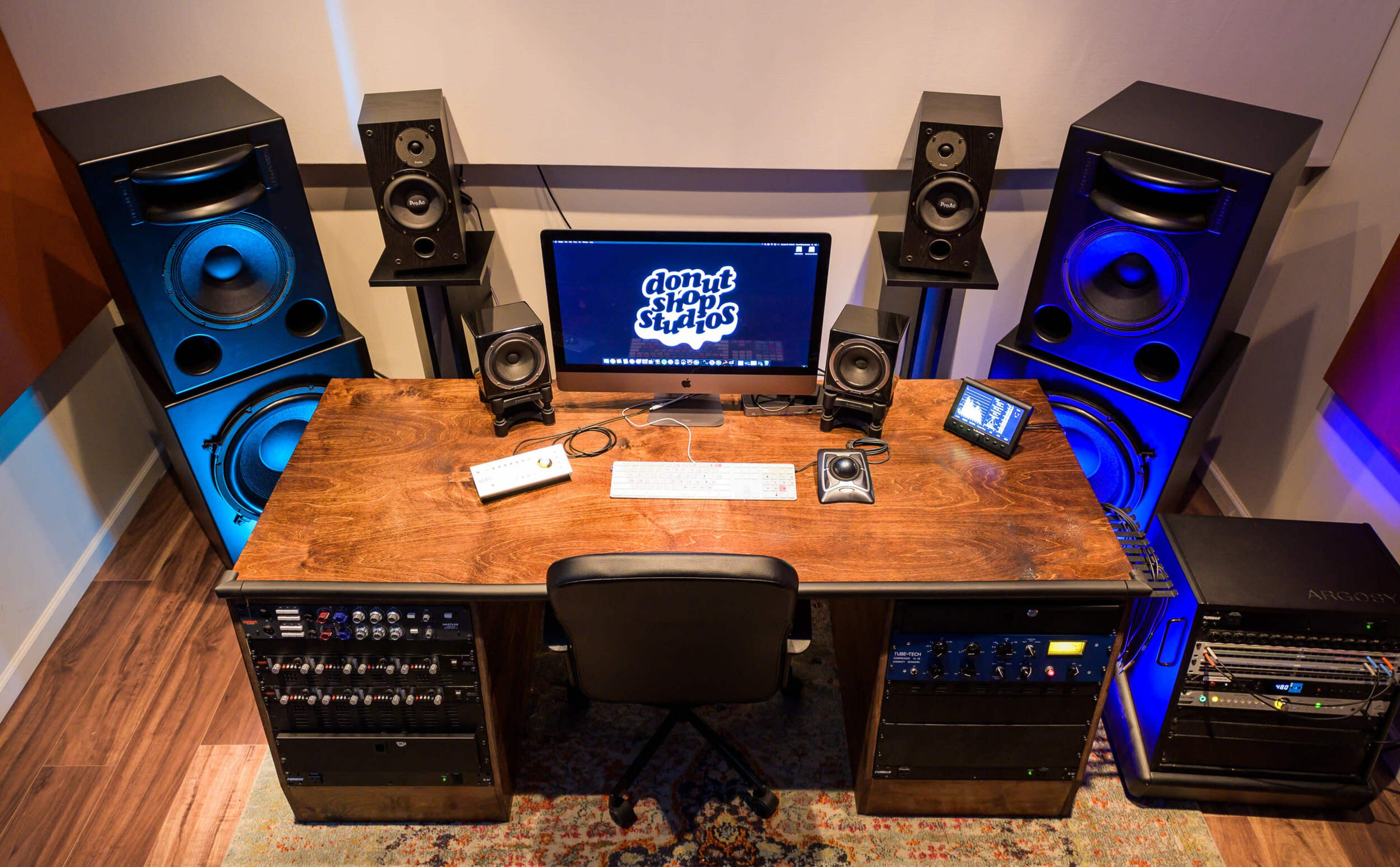 A modern audio production studio featuring a wooden desk with computer monitors, audio equipment, and large speaker stacks on either side.