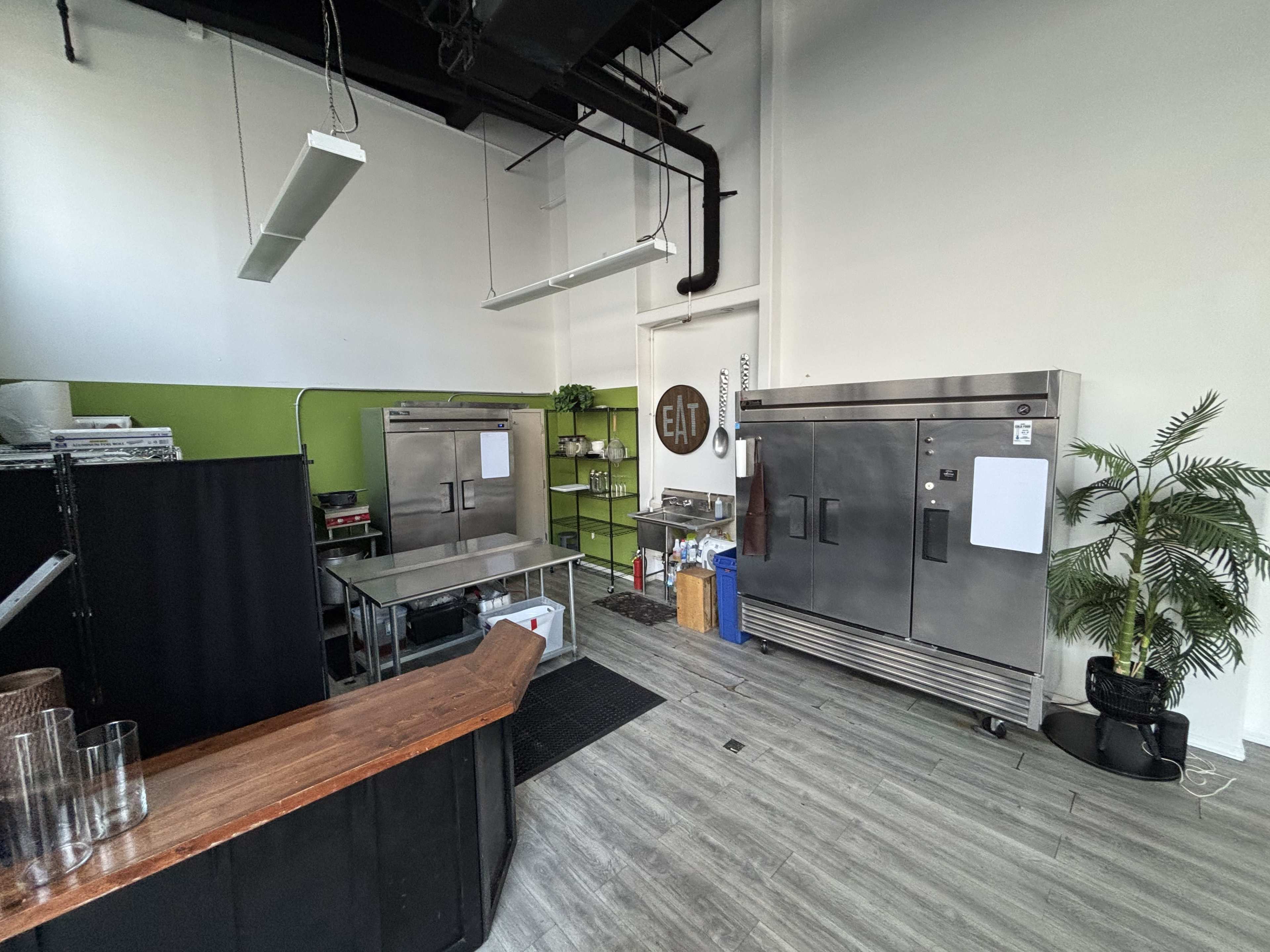 The image shows a modern kitchen space featuring stainless steel refrigerators, a preparation table, and a green accent wall.