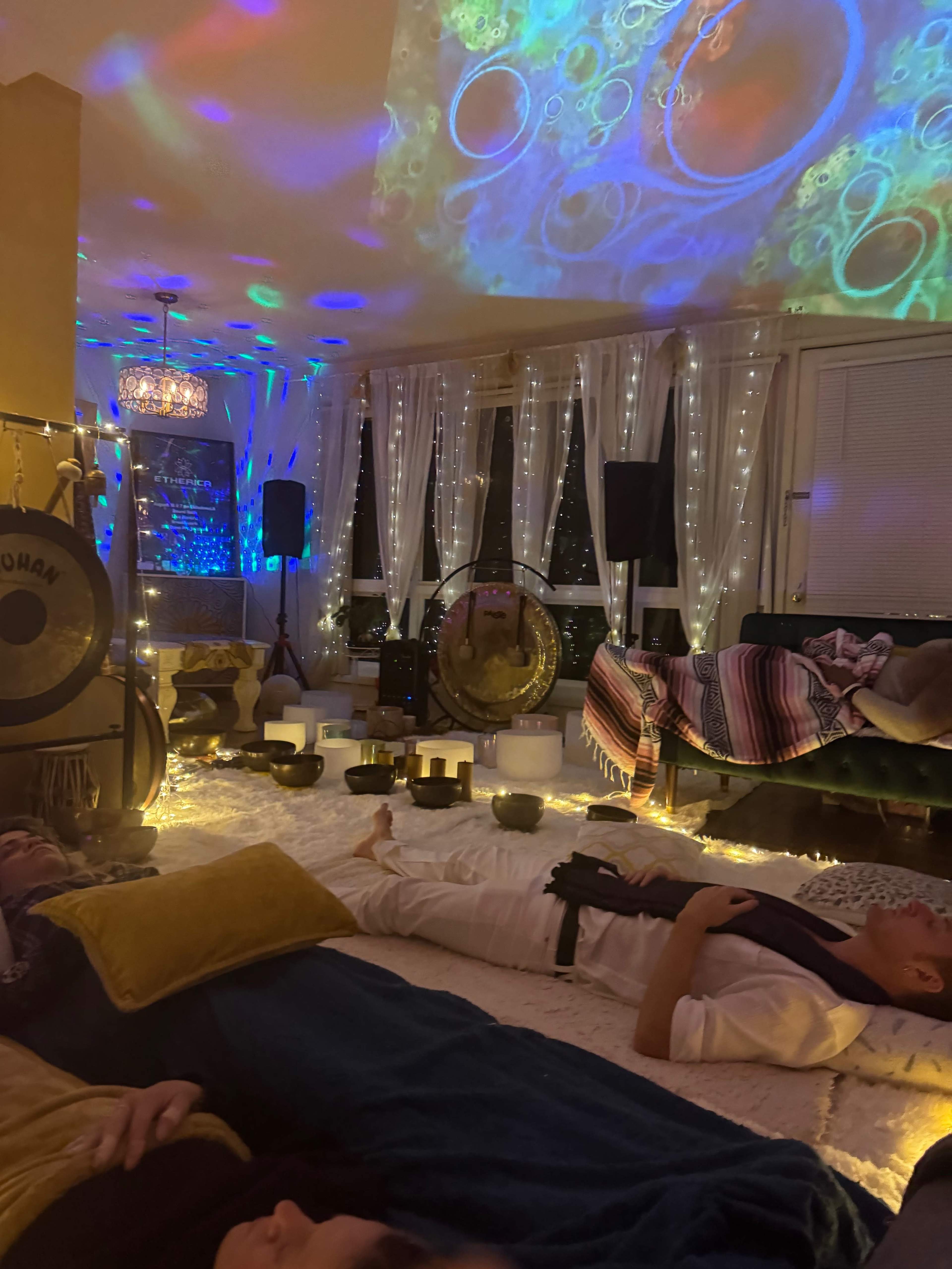 A dimly lit room adorned with colorful projections and string lights, featuring people lying on mats surrounded by musical instruments and soft decor.