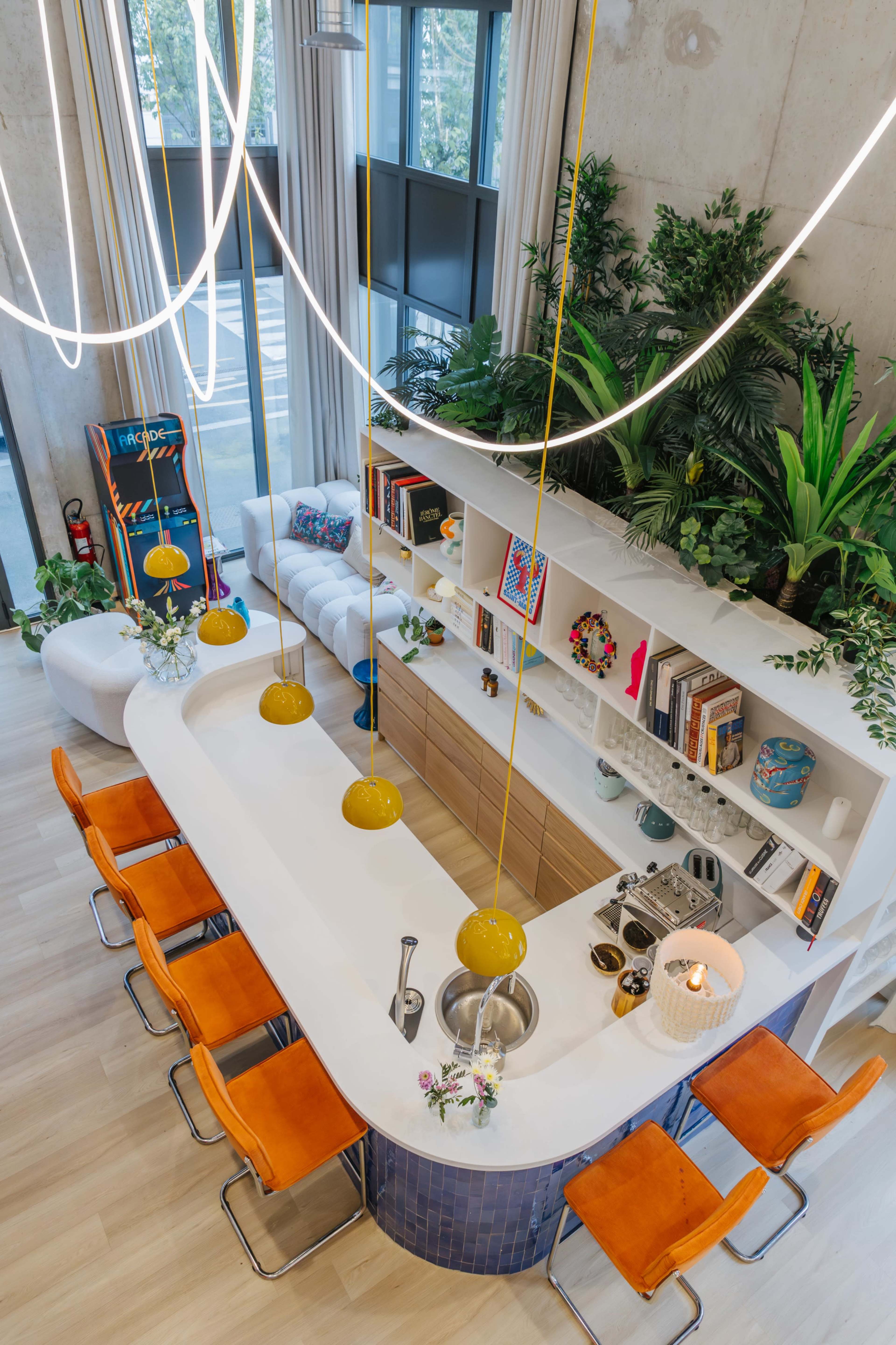 The image shows a modern kitchen with a curved bar counter, orange stools, and pendant lights, surrounded by greenery and decorative items on shelves.