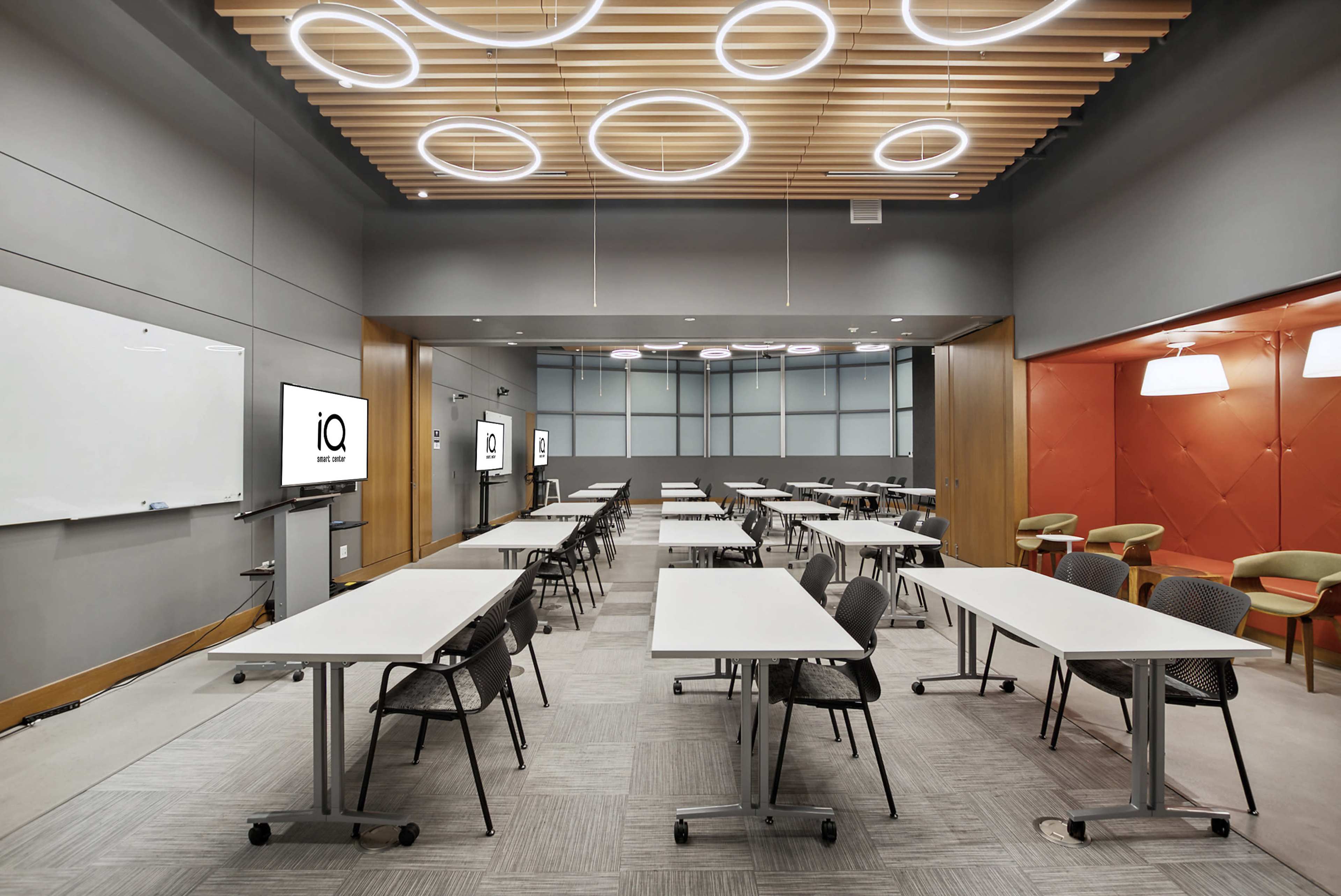 A modern conference room features several rows of tables and chairs, illuminated by circular ceiling lights and equipped with presentation screens.