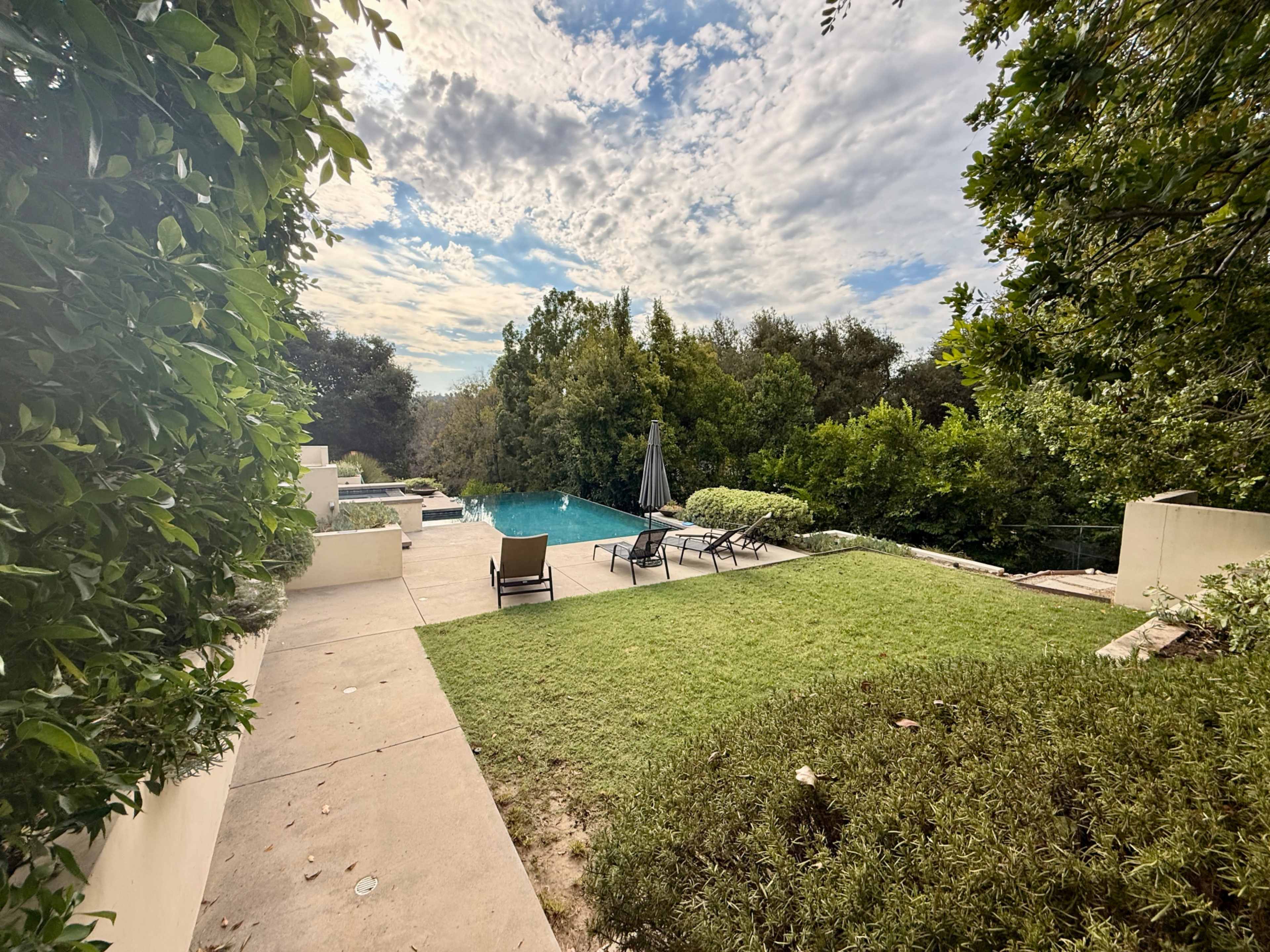 The image shows a serene backyard with a swimming pool surrounded by greenery, loungers, and a patio area under a partly cloudy sky.