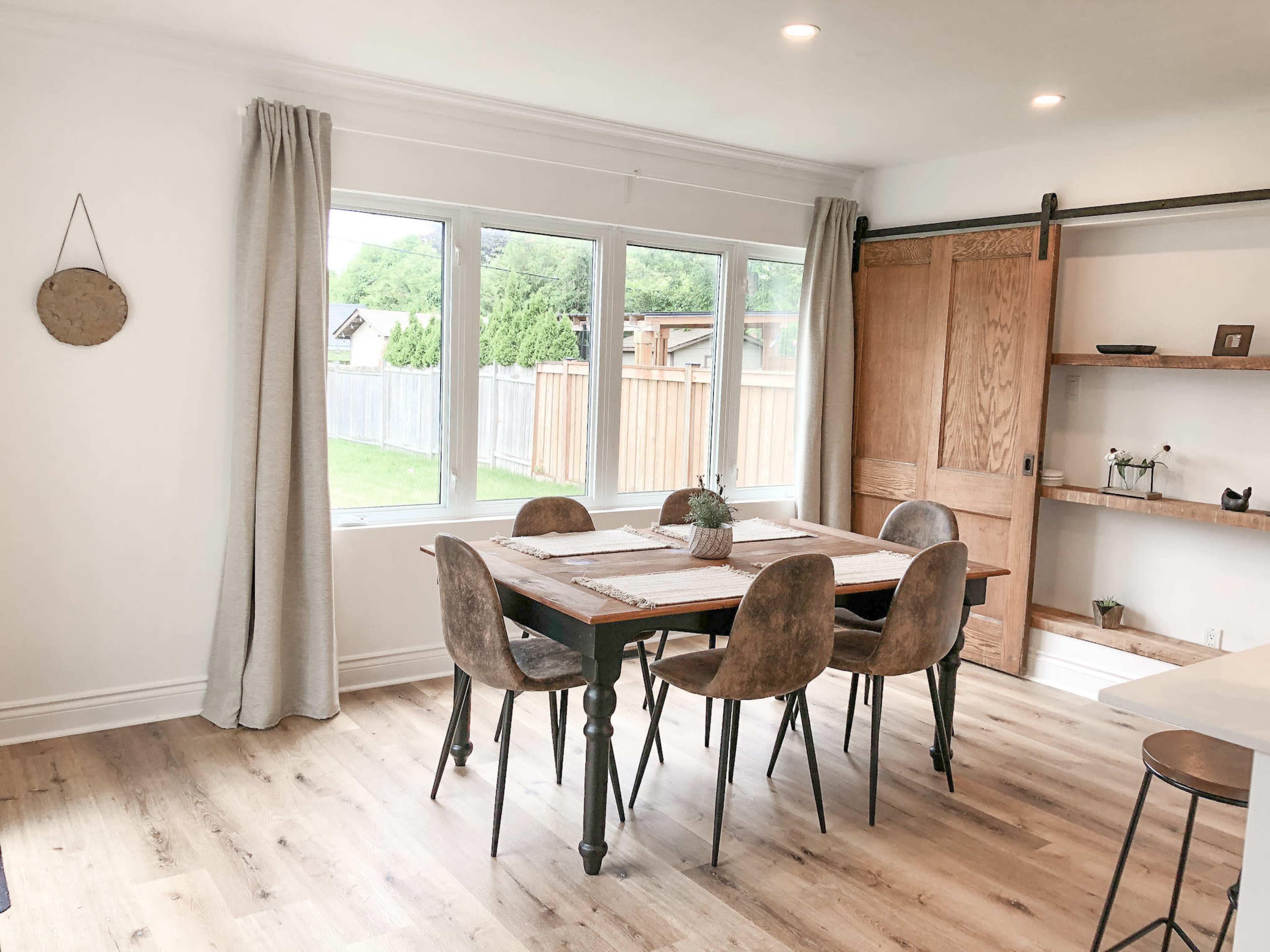 A dining area features a wooden table surrounded by chairs, with large windows overlooking a grassy yard and a sliding barn door on one side.