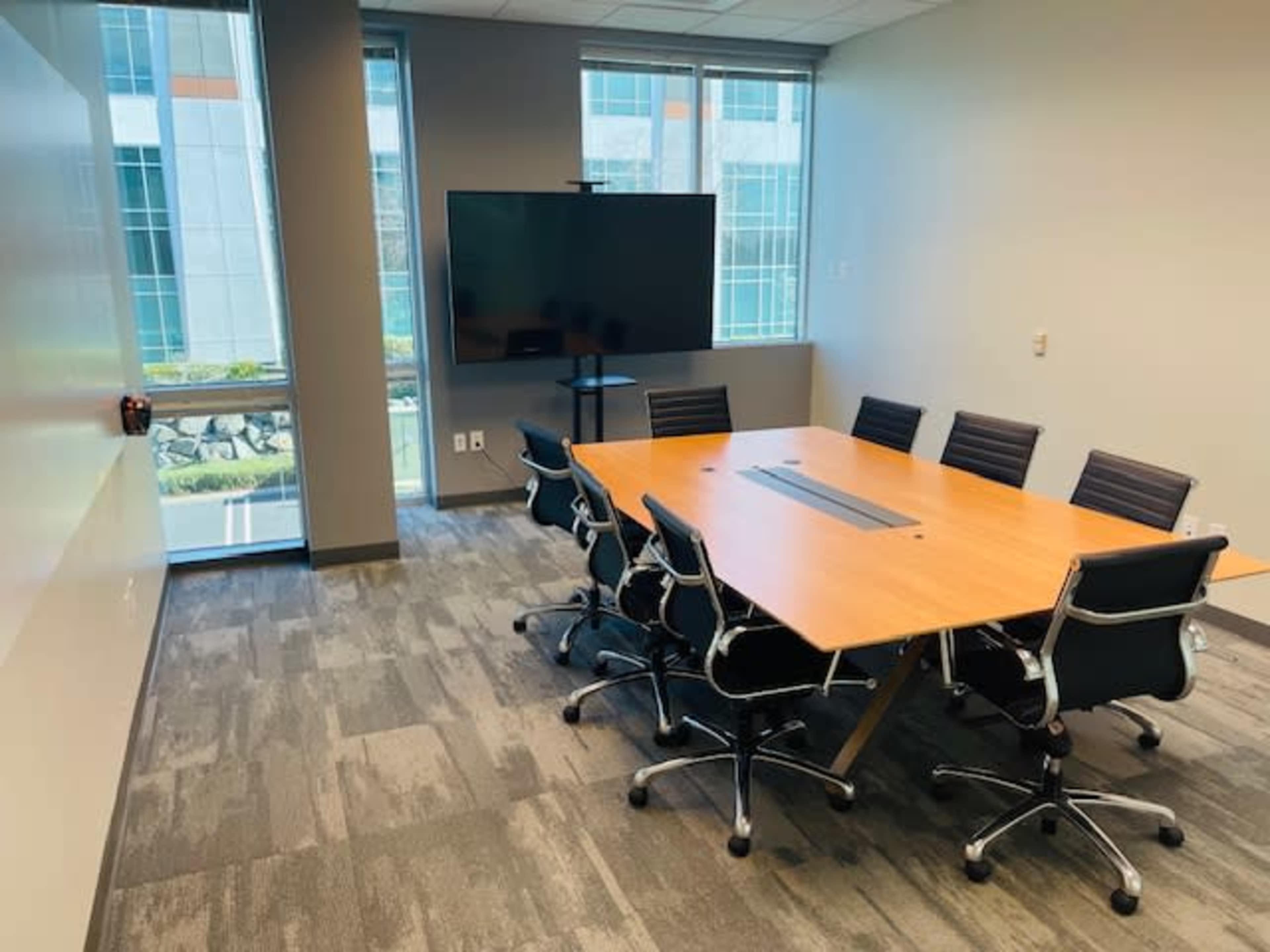 A modern conference room features a large wooden table surrounded by eight black chairs, with a wall-mounted screen and windows offering a view outside.