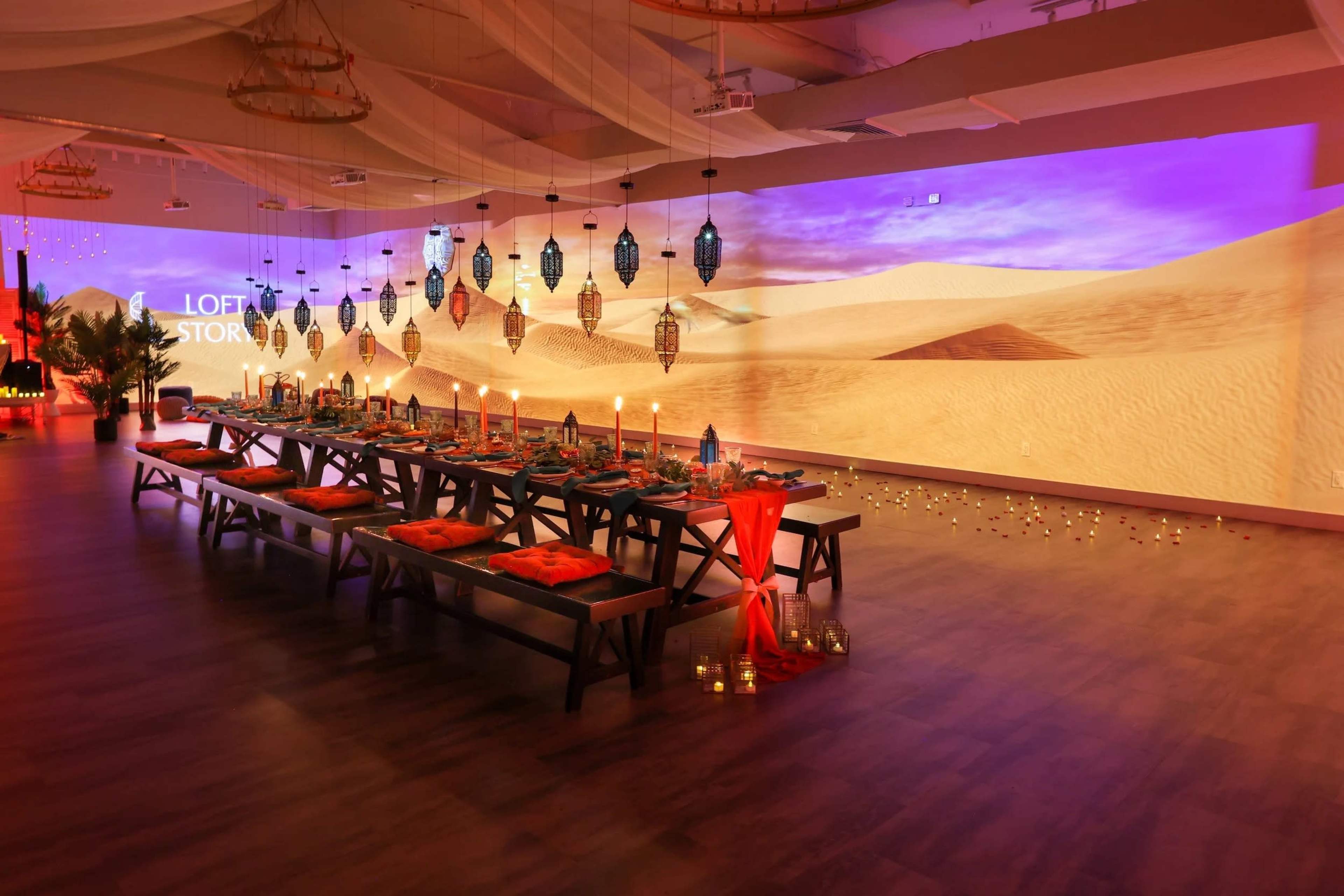 The image shows a beautifully arranged dining setup with a long table adorned with decorations and surrounded by lanterns, set in a space featuring a desert-themed projected backdrop.