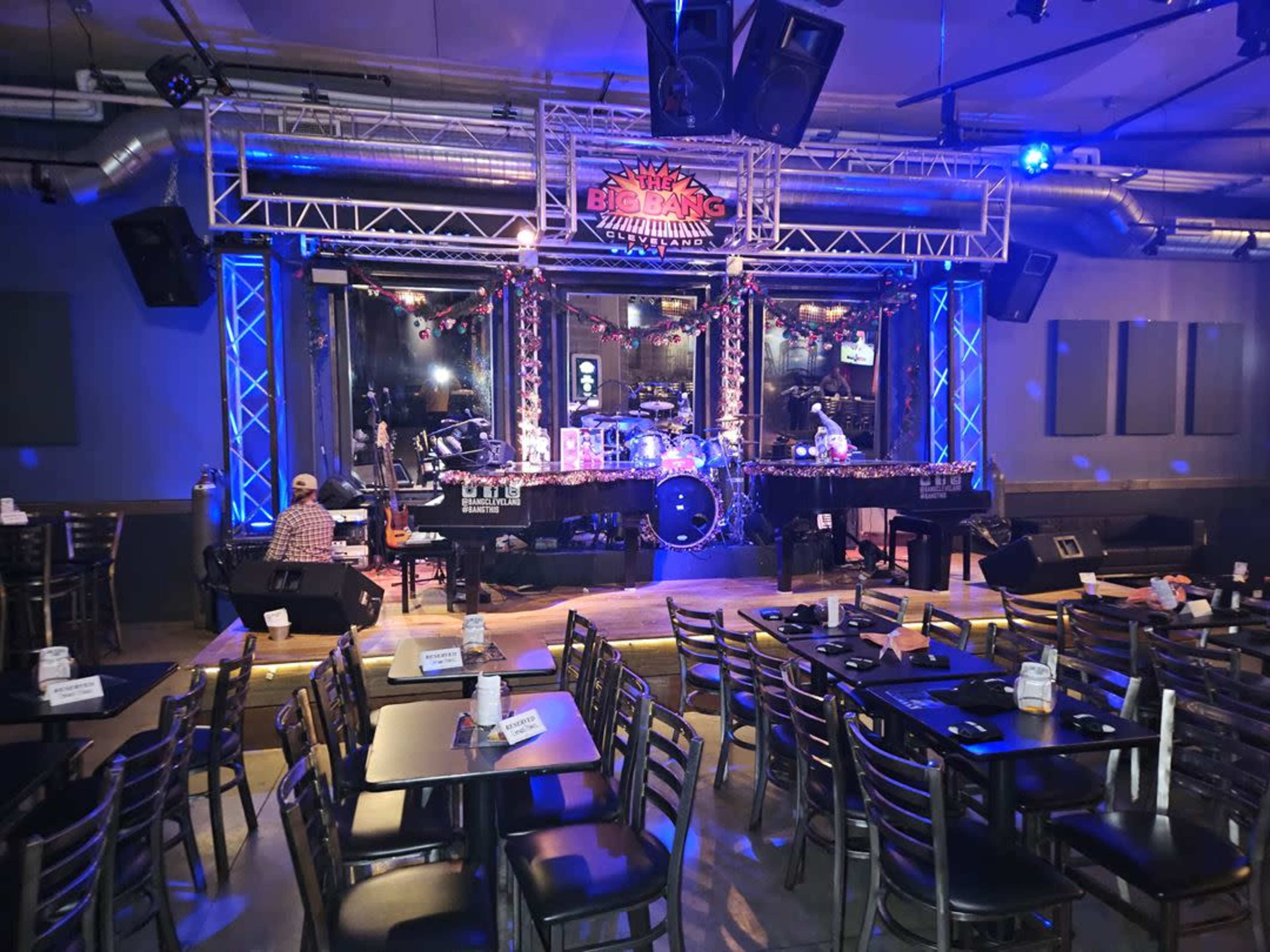 A live music venue features a stage with instruments set up under bright lights, surrounded by tables and chairs.
