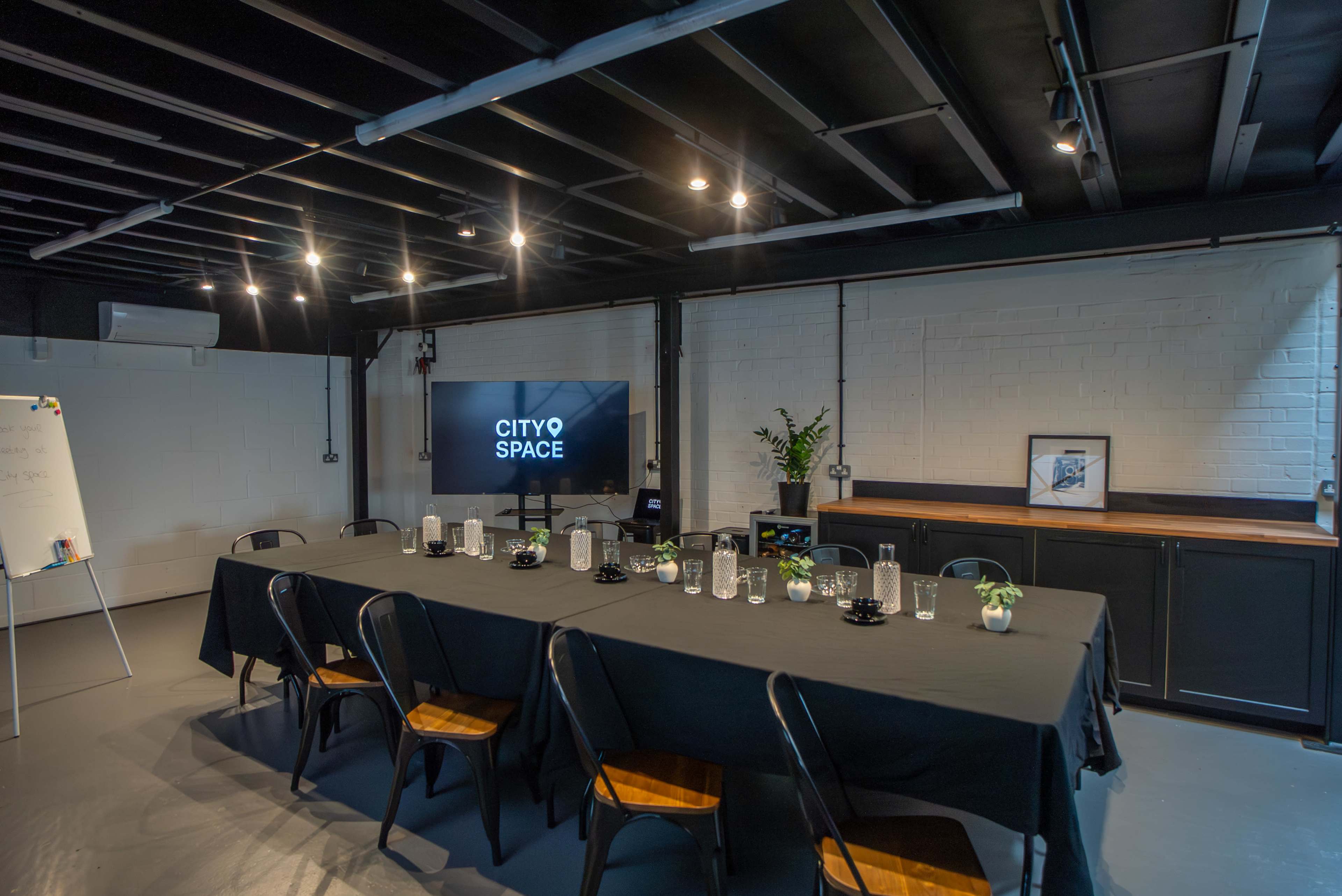 The image shows a modern conference room with a long black table set for a meeting, equipped with a television screen displaying "CITY SPACE" and a whiteboard nearby.