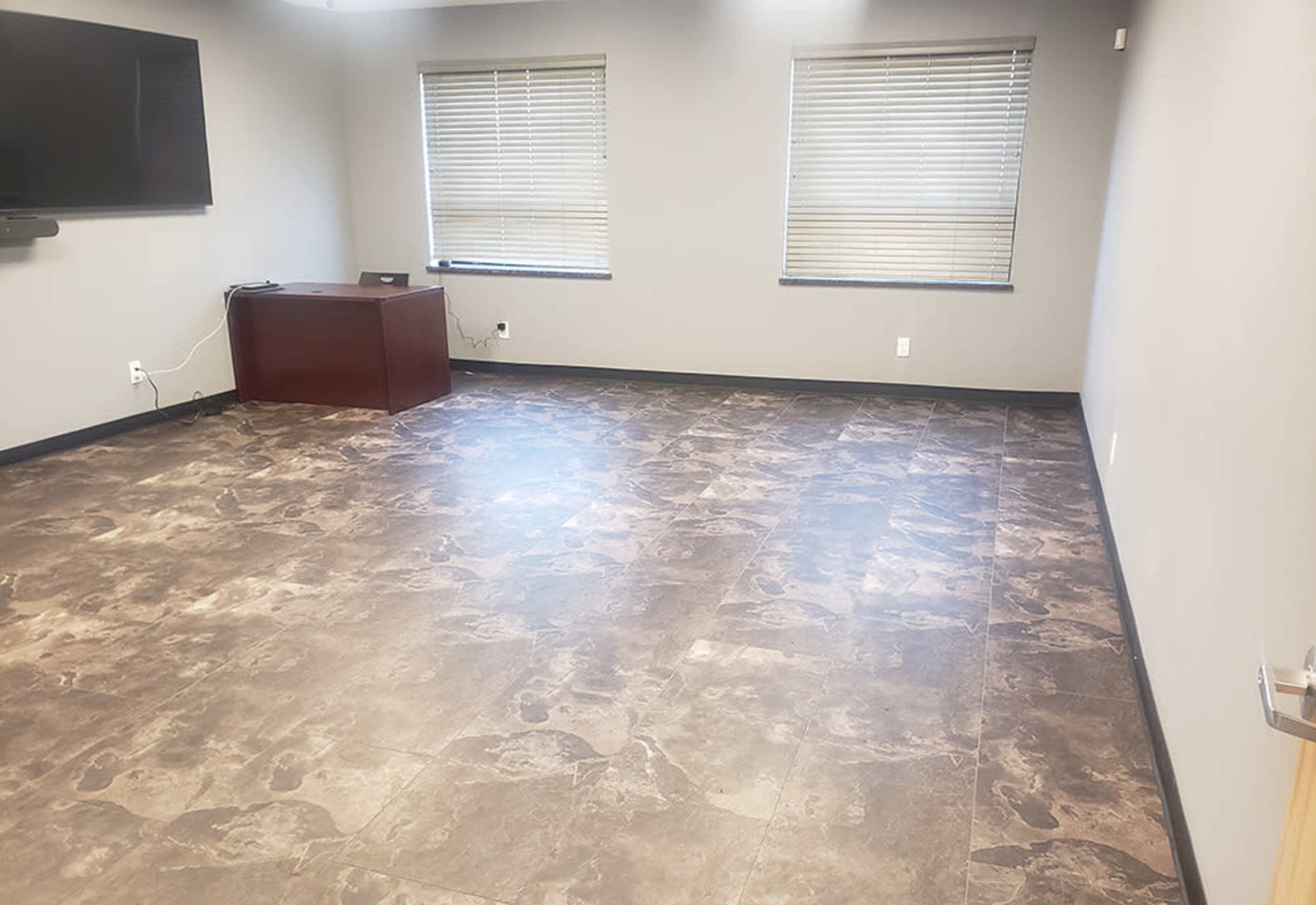 The image shows an empty room with a smooth, patterned floor, two windows with blinds, a wall-mounted television, and a dark wooden cabinet.