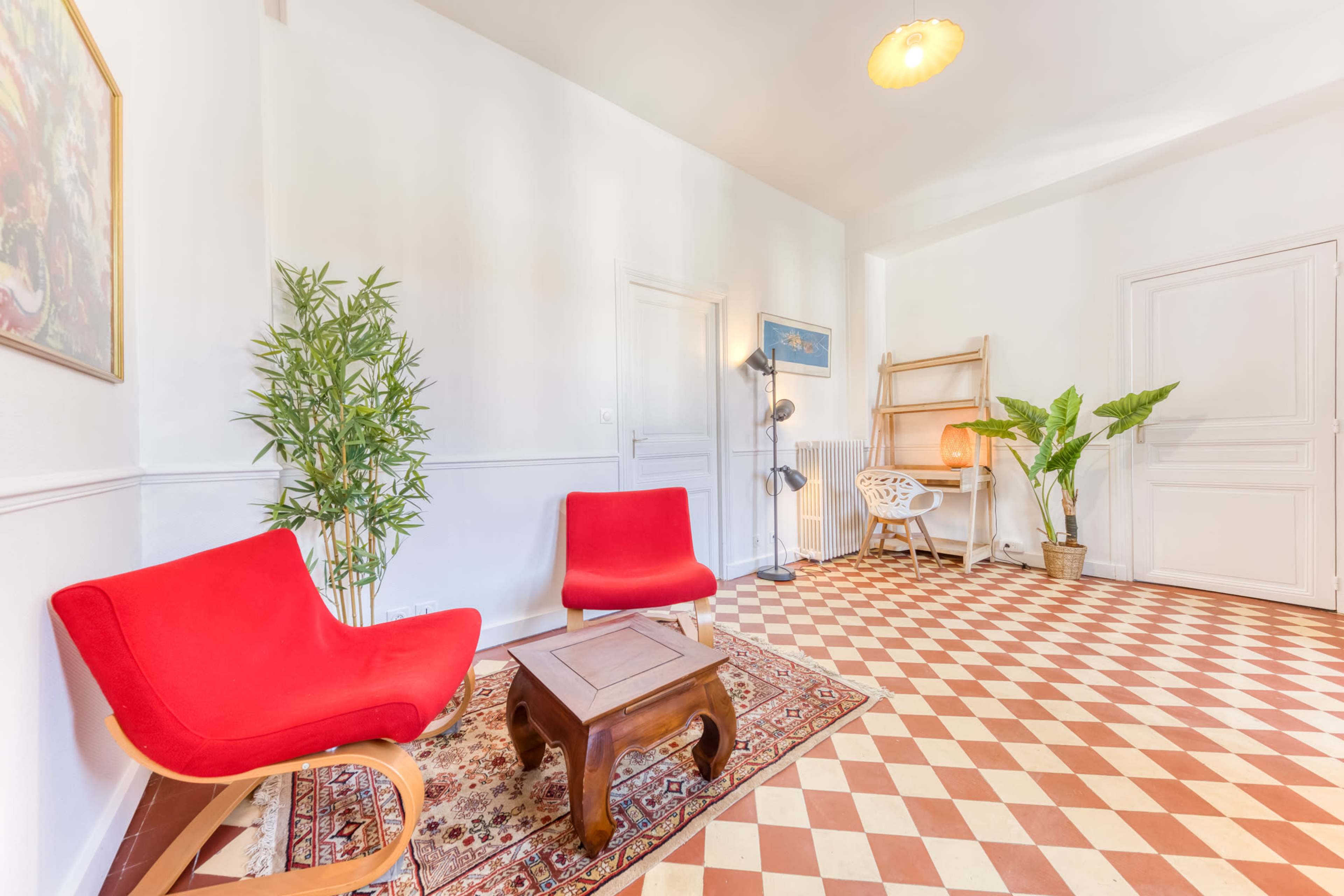 The image shows a bright, minimalistic room with red chairs, a small wooden table, plants, and a patterned tile floor.