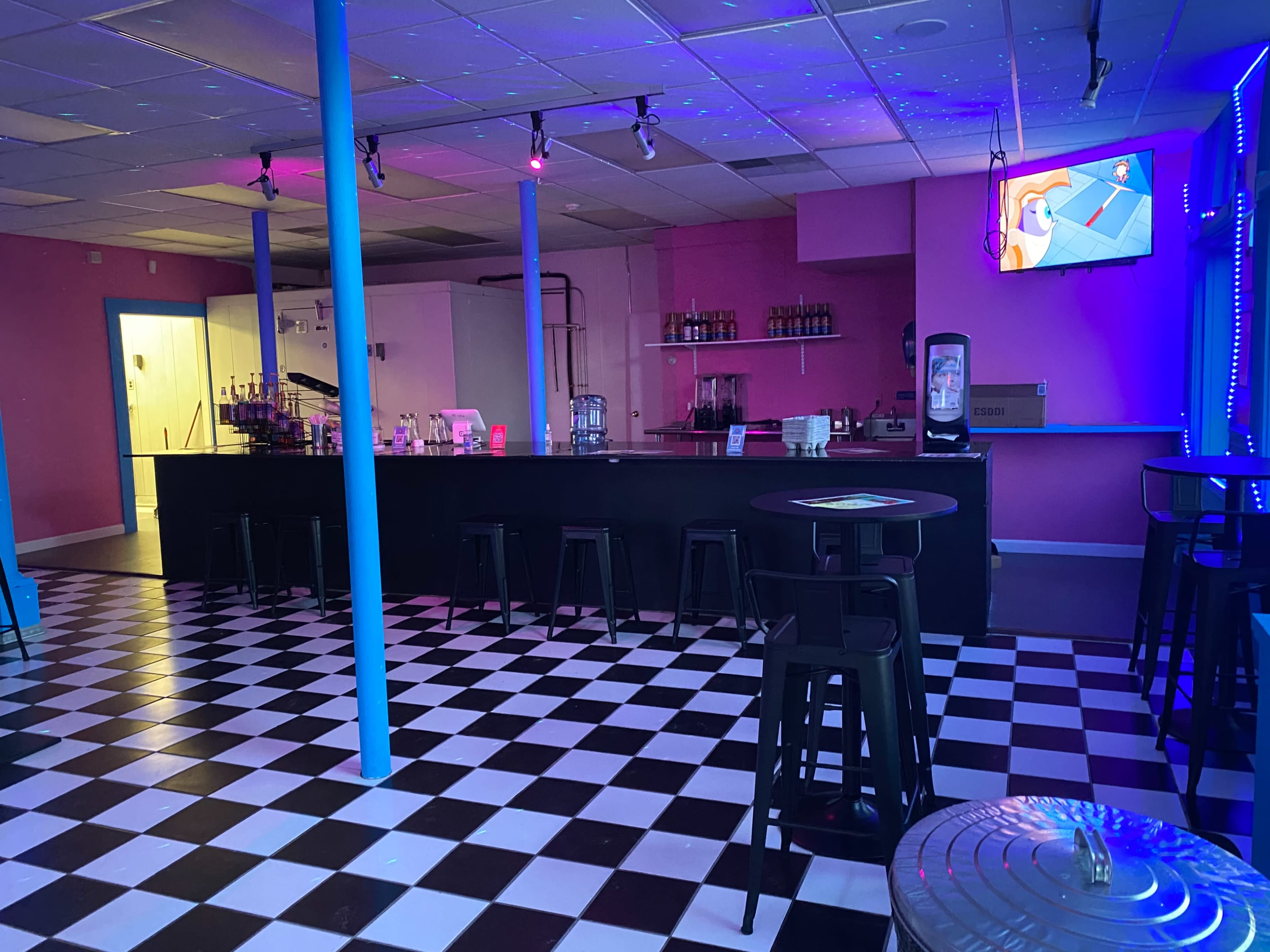 A colorful bar area with a checkered black and white floor, a pink wall, and a TV mounted on the right displaying a vibrant scene.