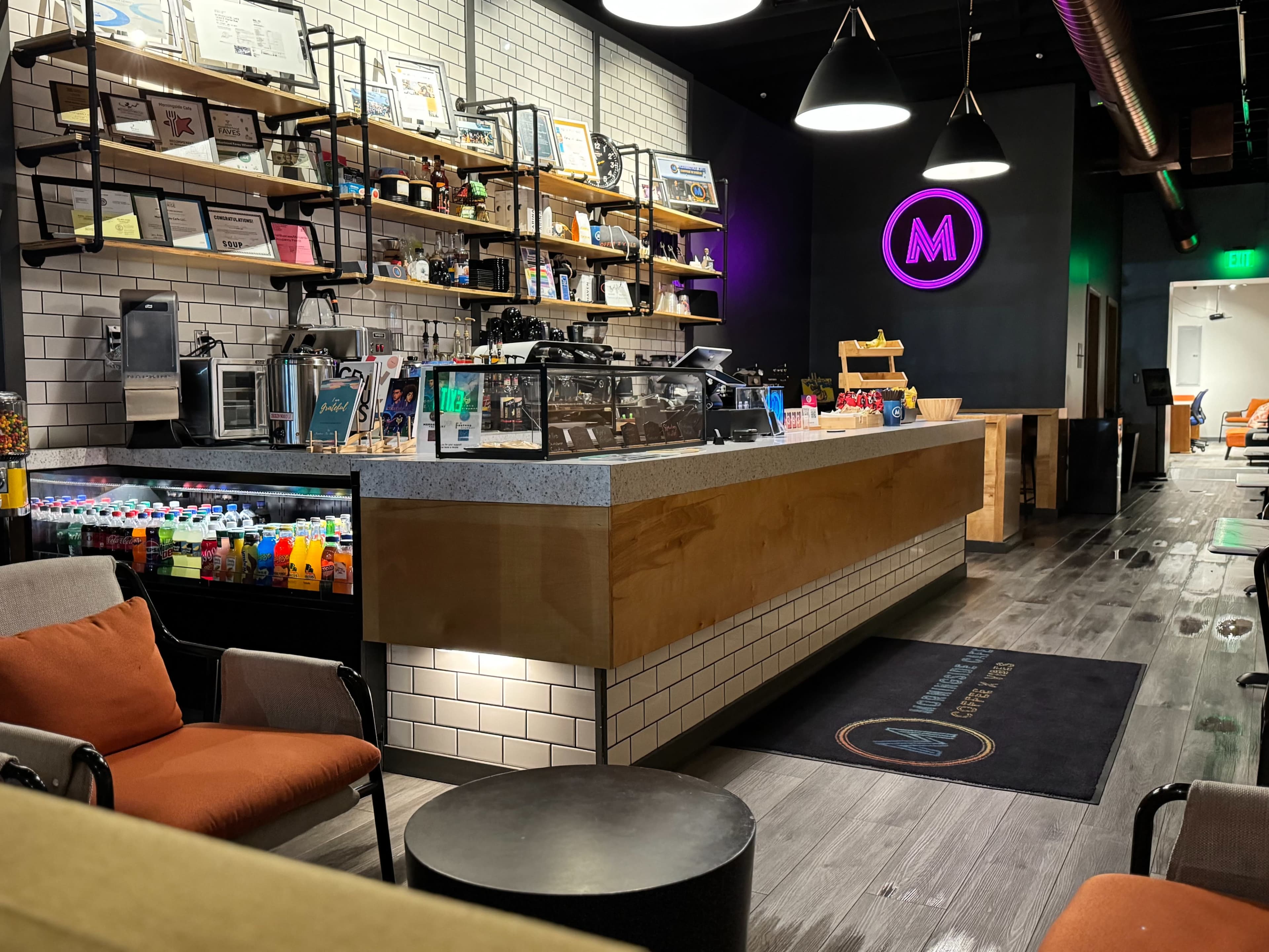 The image shows a modern cafe interior featuring a counter with shelves displaying various items, seating areas with orange chairs, and a neon sign.