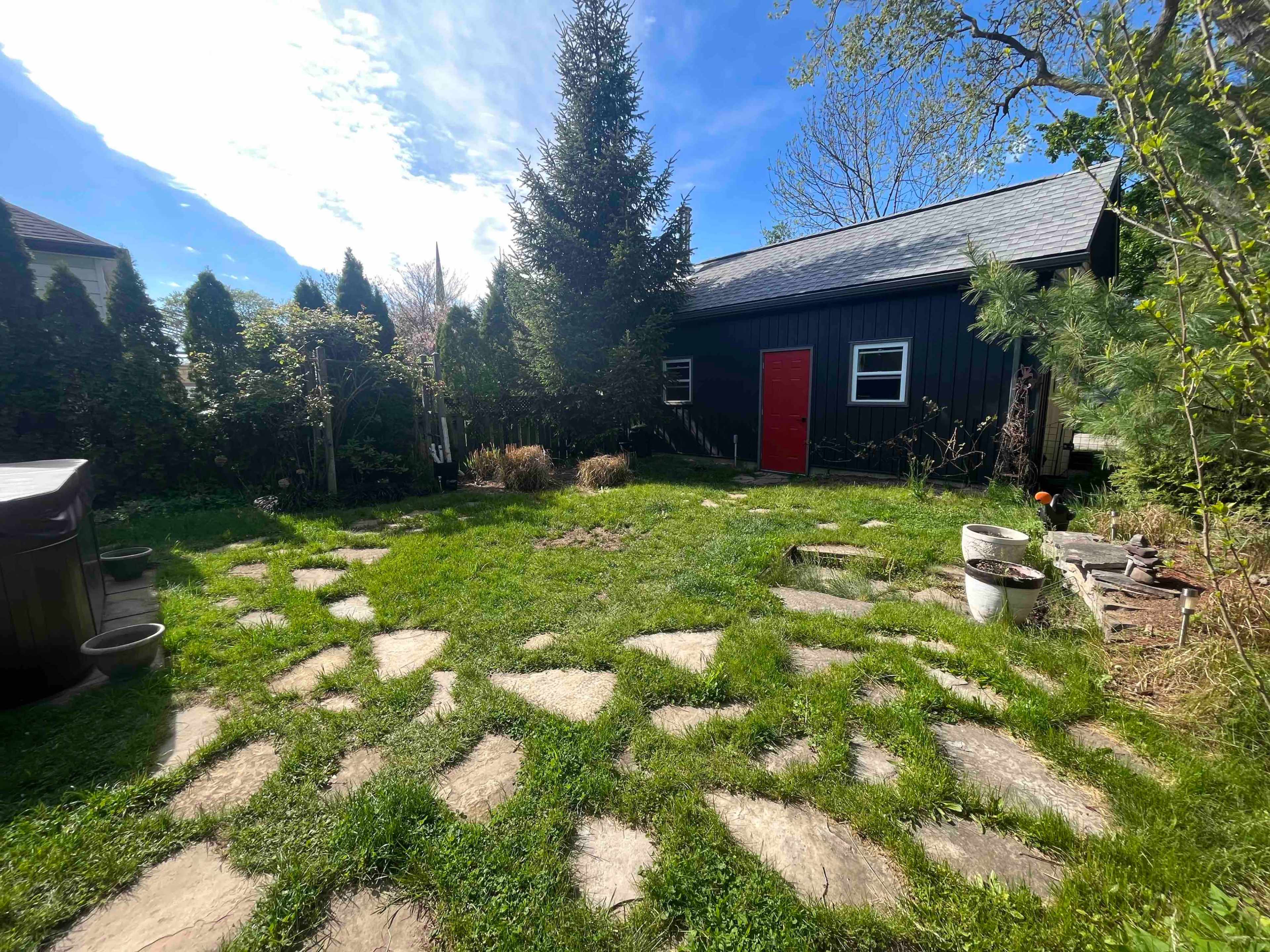 A garden area features a stone pathway leading to a black shed with a red door, surrounded by greenery and trees.