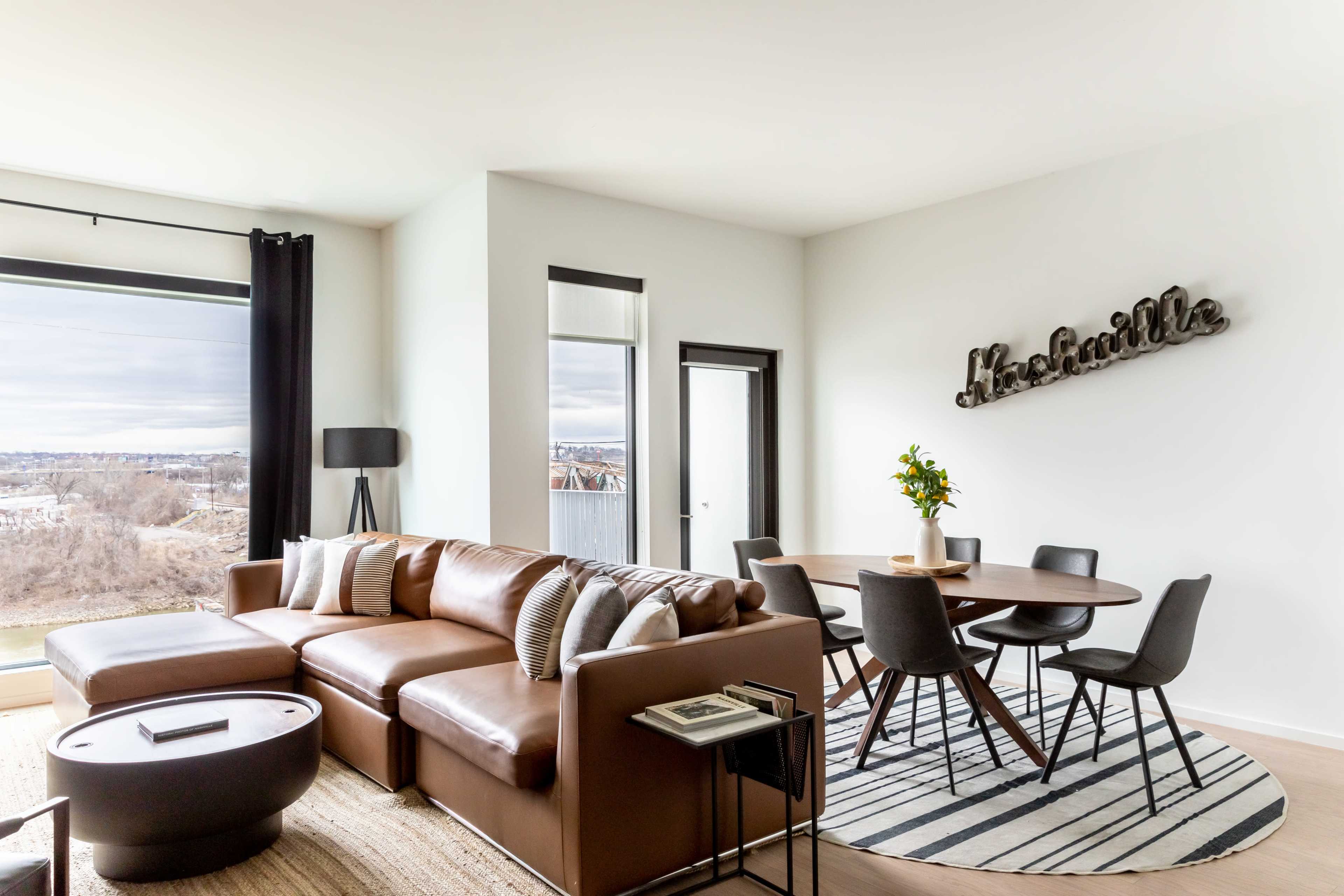 A modern living room and dining area with a brown sectional sofa, round dining table, and large windows overlooking a scenic view.