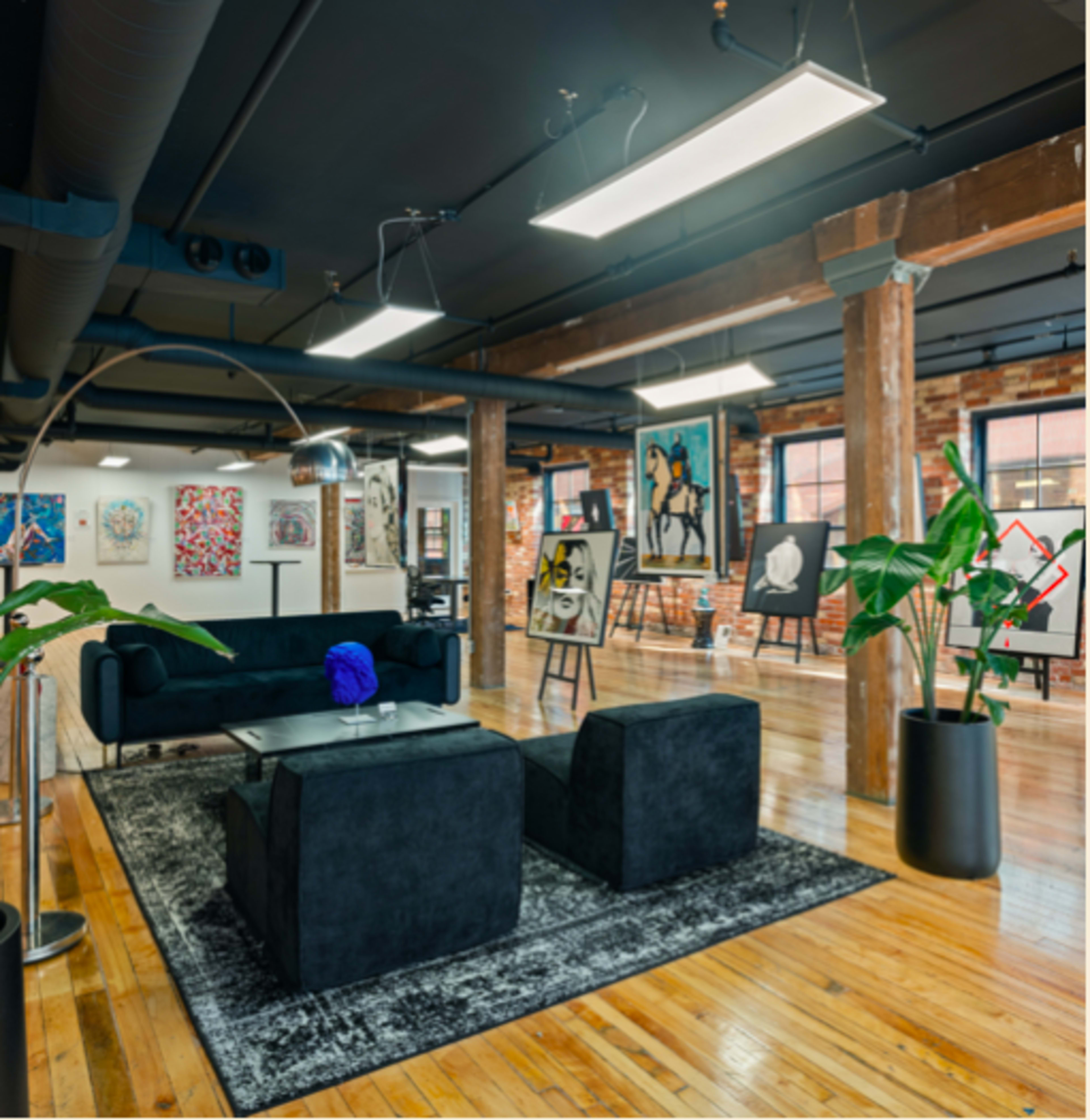 The image shows a modern art gallery with a black sectional sofa, two matching chairs, and various artworks displayed on the walls.