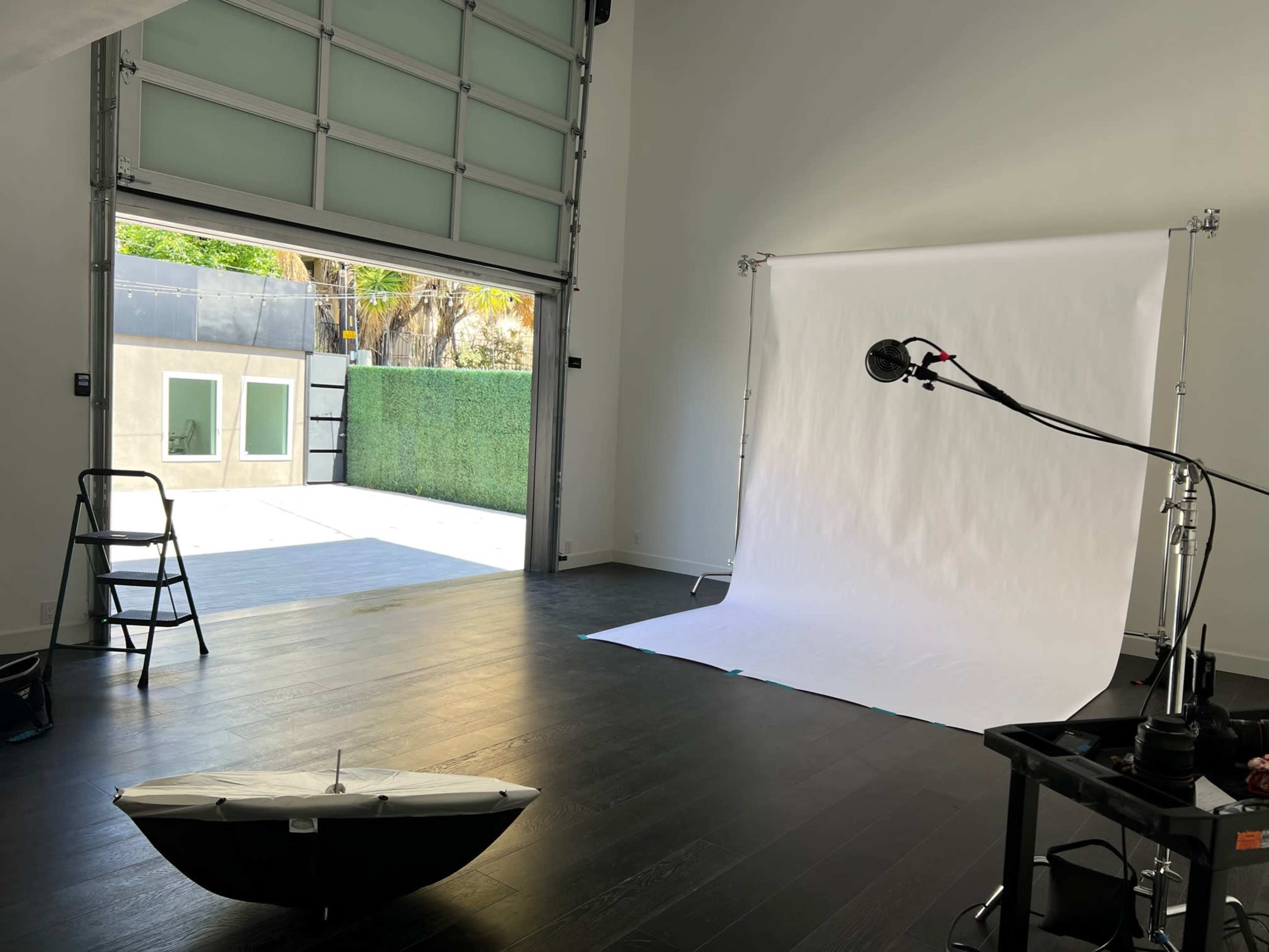 A spacious photography studio with a backdrop setup, a microphone stand, a ladder, and an open garage door revealing an outdoor area.