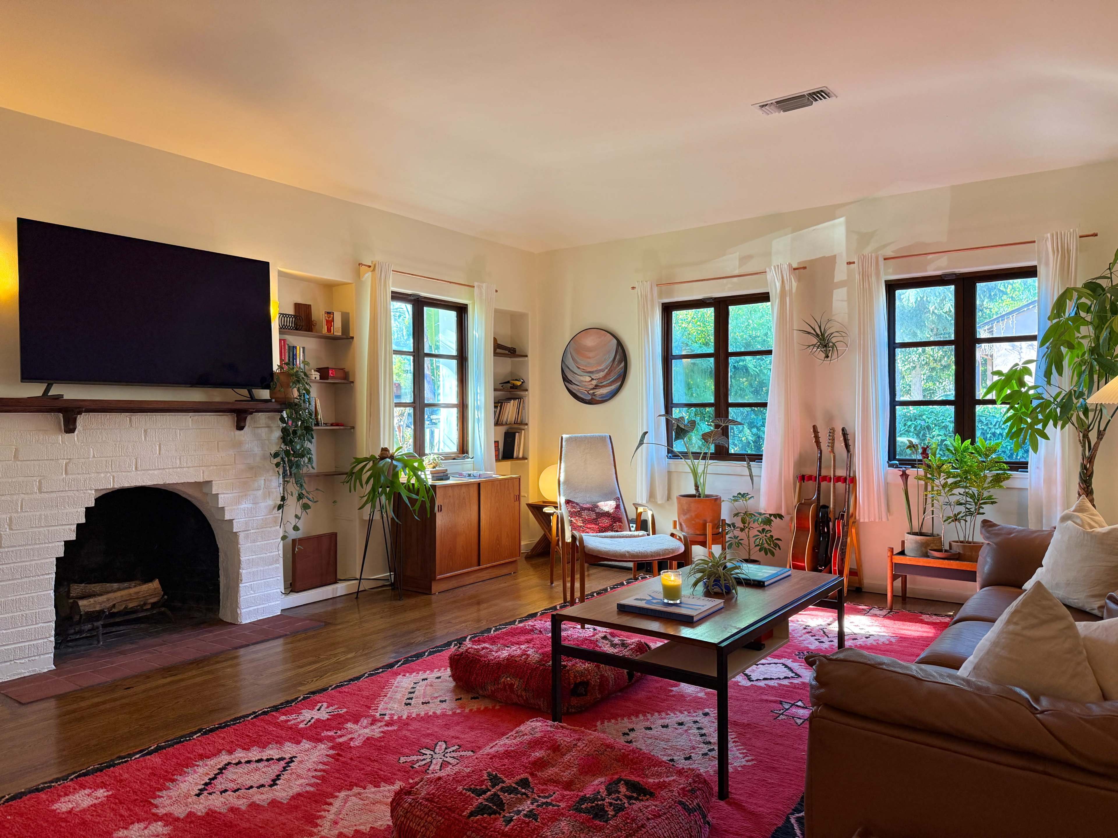 The living room features a cozy arrangement with a large television mounted on the wall, a fireplace, comfortable seating, and multiple plants in front of bright windows.