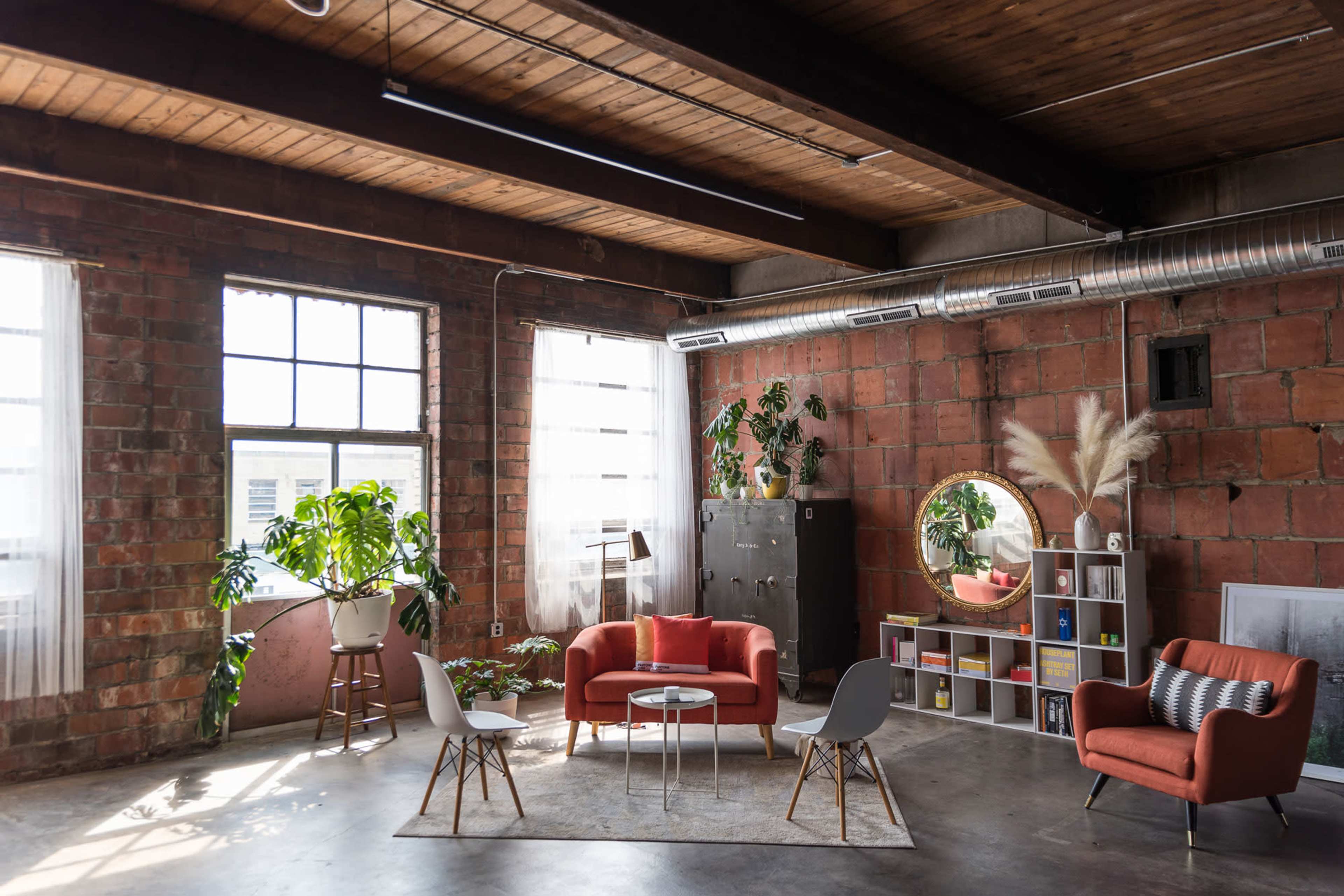 The image shows a modern, industrial-style living space with exposed brick walls, large windows, and contemporary furniture arrangements including a red sofa and white chairs.