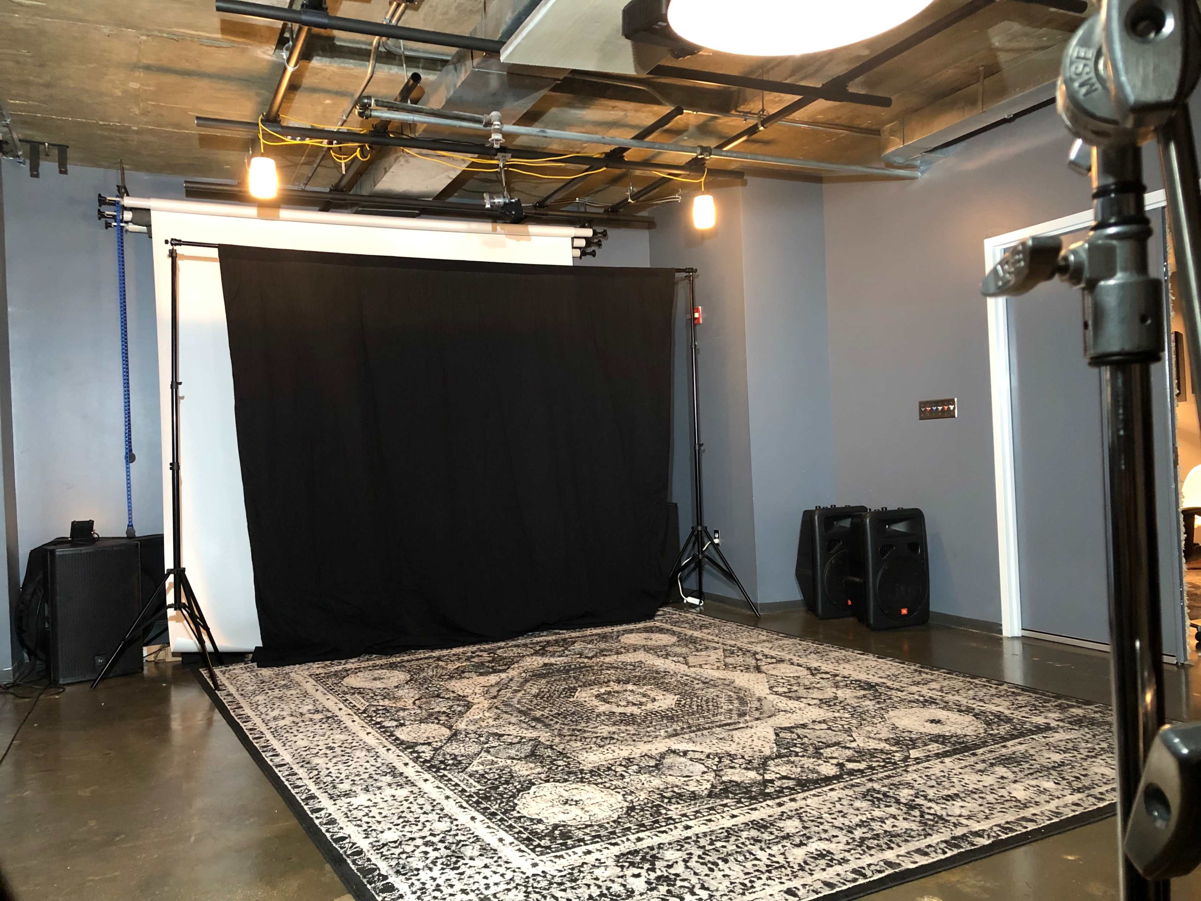 A photography studio featuring a black backdrop, a patterned rug, and lighting fixtures on the ceiling.