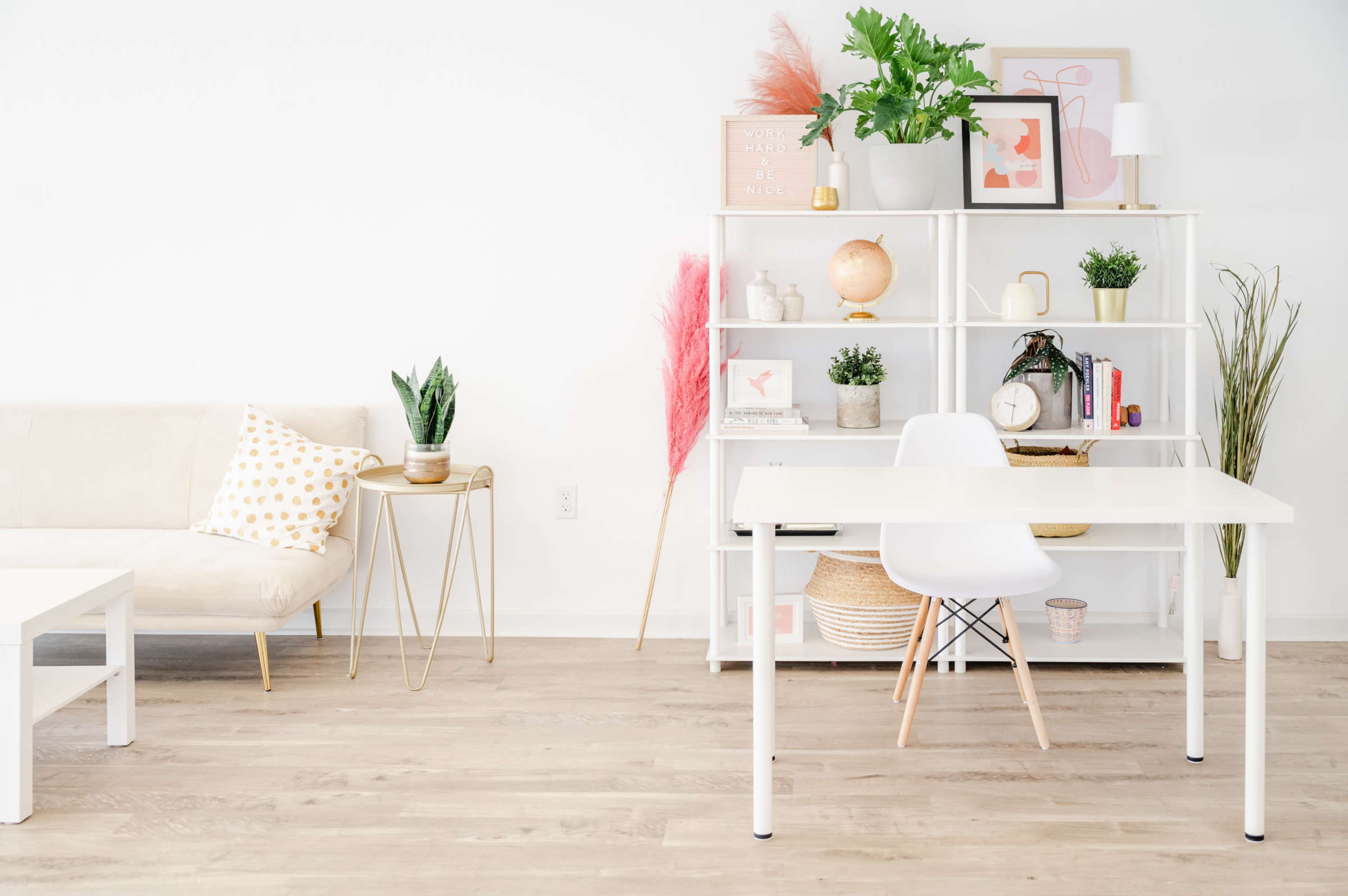 A bright and minimalistic room features a white desk, a beige couch with decorative pillows, and a modern shelving unit adorned with plants and various decor items.
