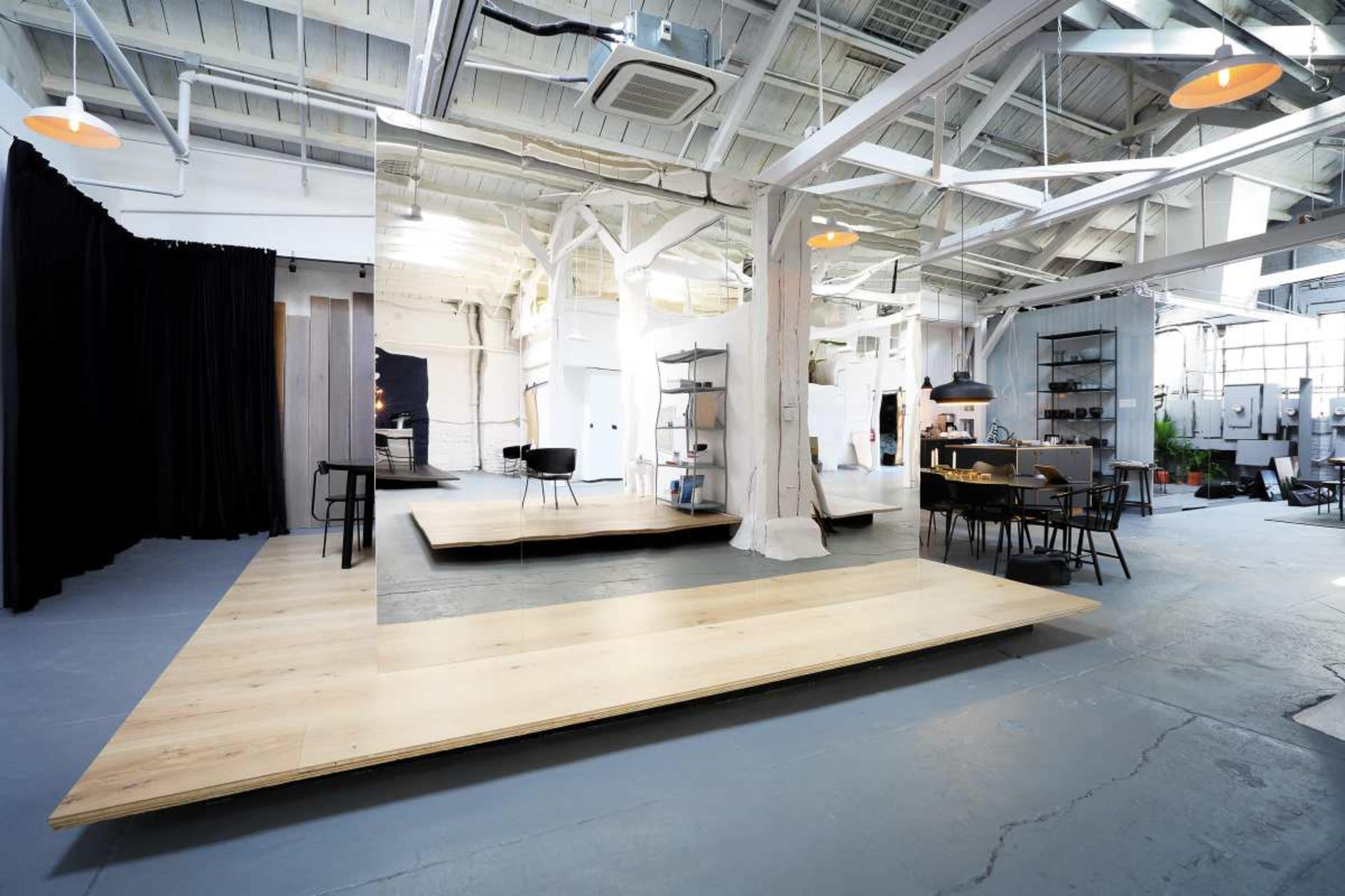 The image shows an industrial-style open space featuring wooden platforms, a black curtain, and various furniture arrangements, set against white walls and a high ceiling.