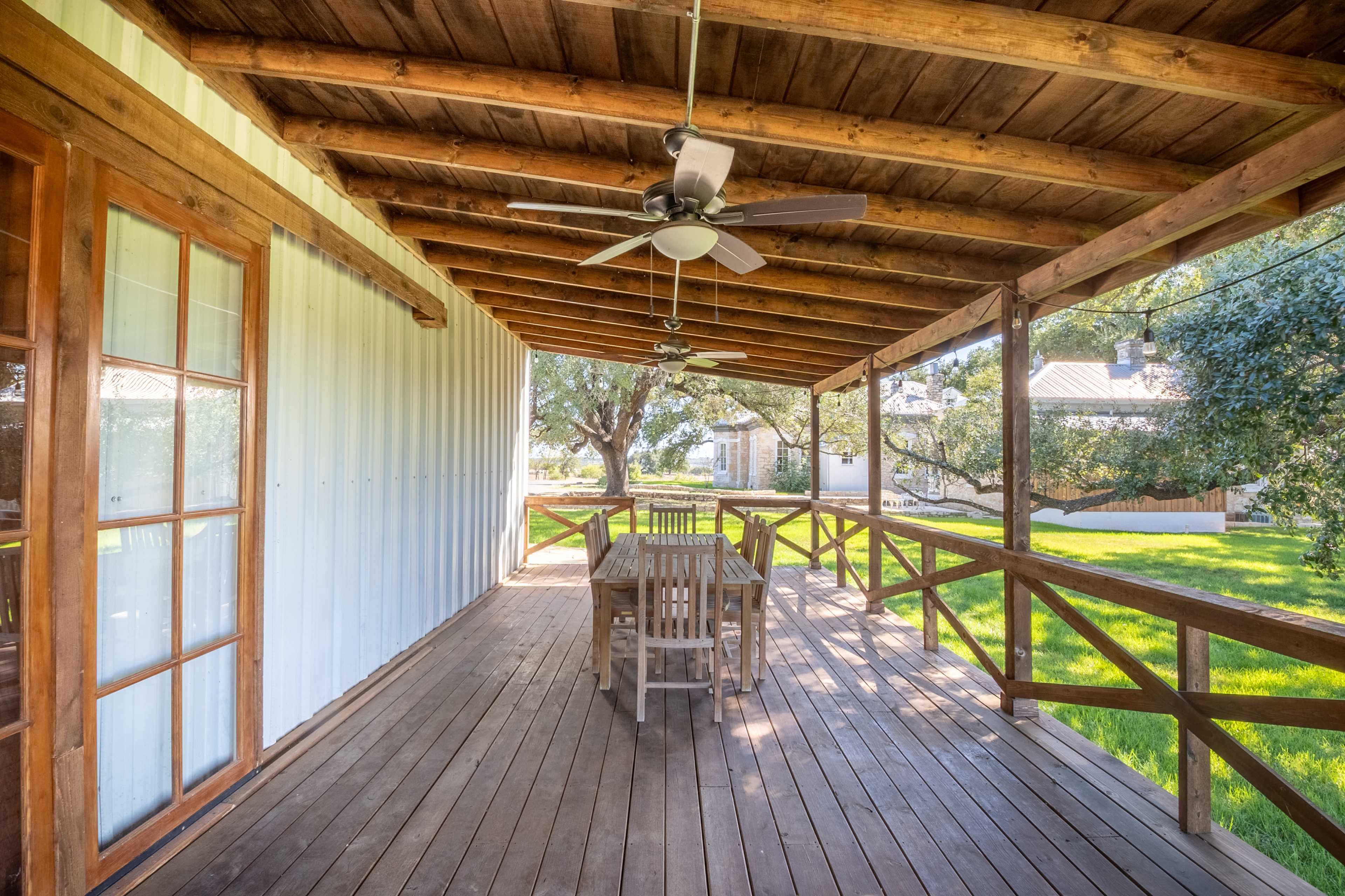 A wooden deck with a table and chairs overlooks a grassy area, featuring ceiling fans and a covered roof.