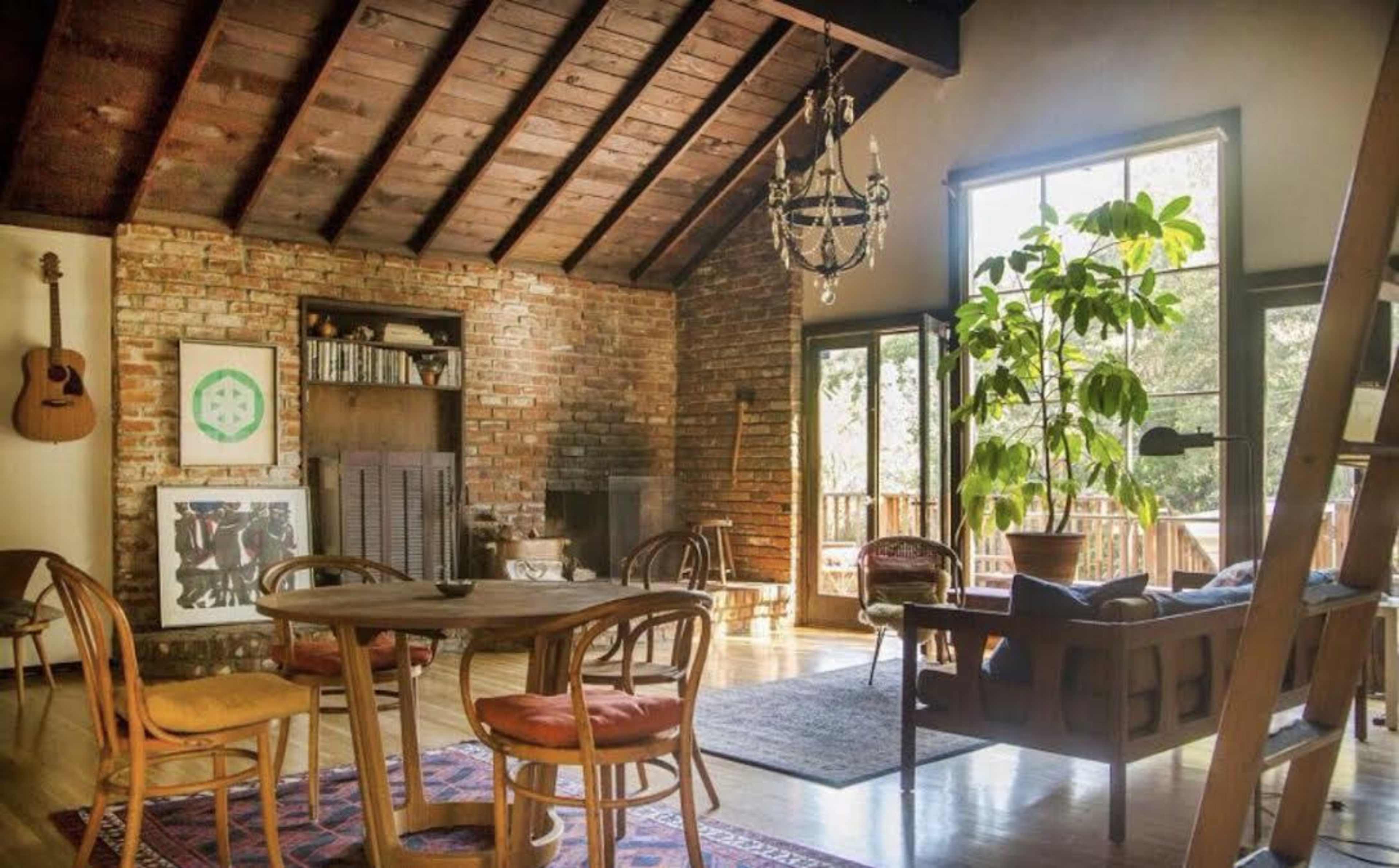 A cozy living room features a wooden ceiling, a brick wall, a dining table, and large windows with natural light streaming in.