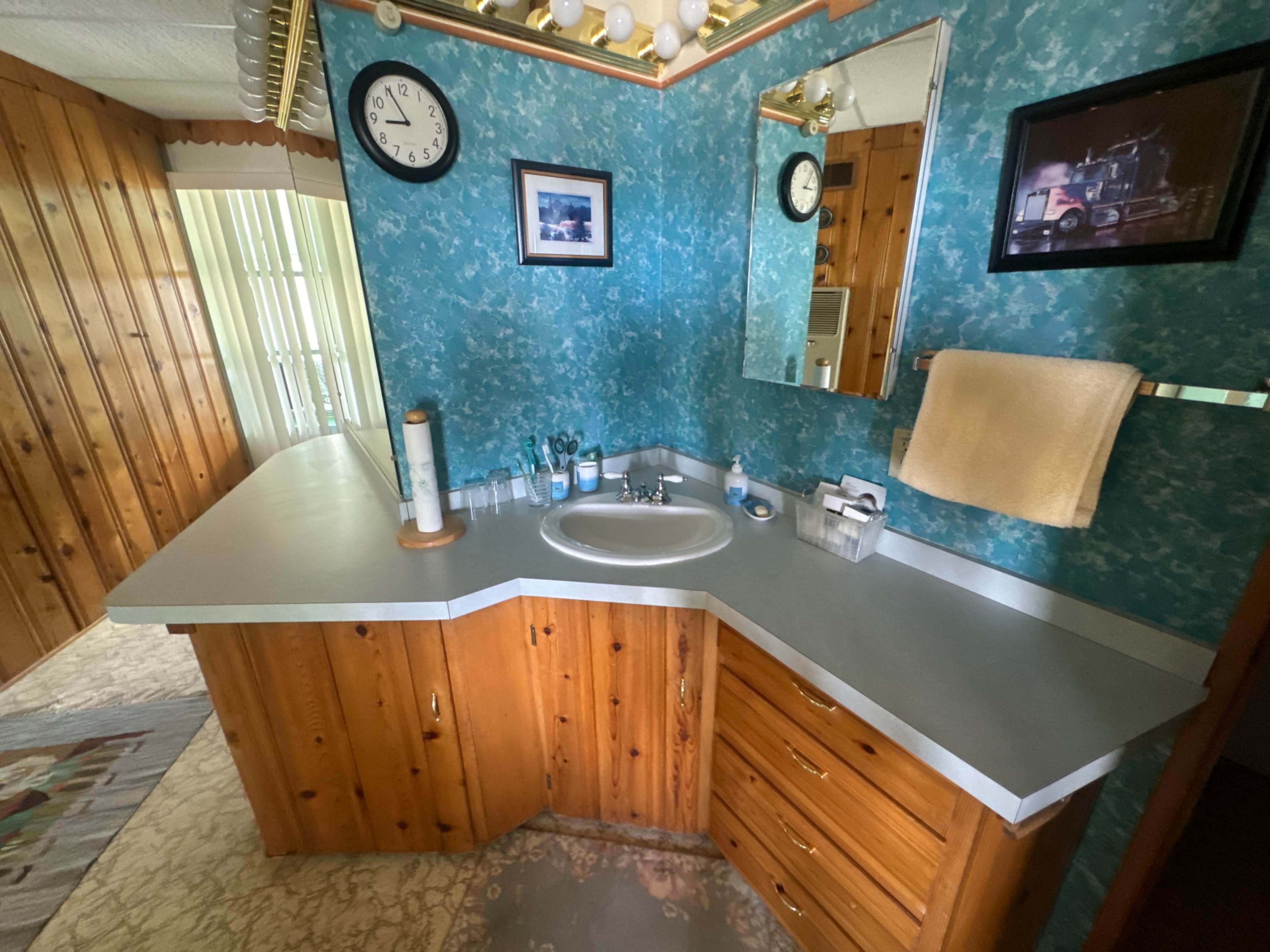 The image shows a bathroom with a triangular sink area featuring wooden cabinets, a mirror, and turquoise wallpaper.