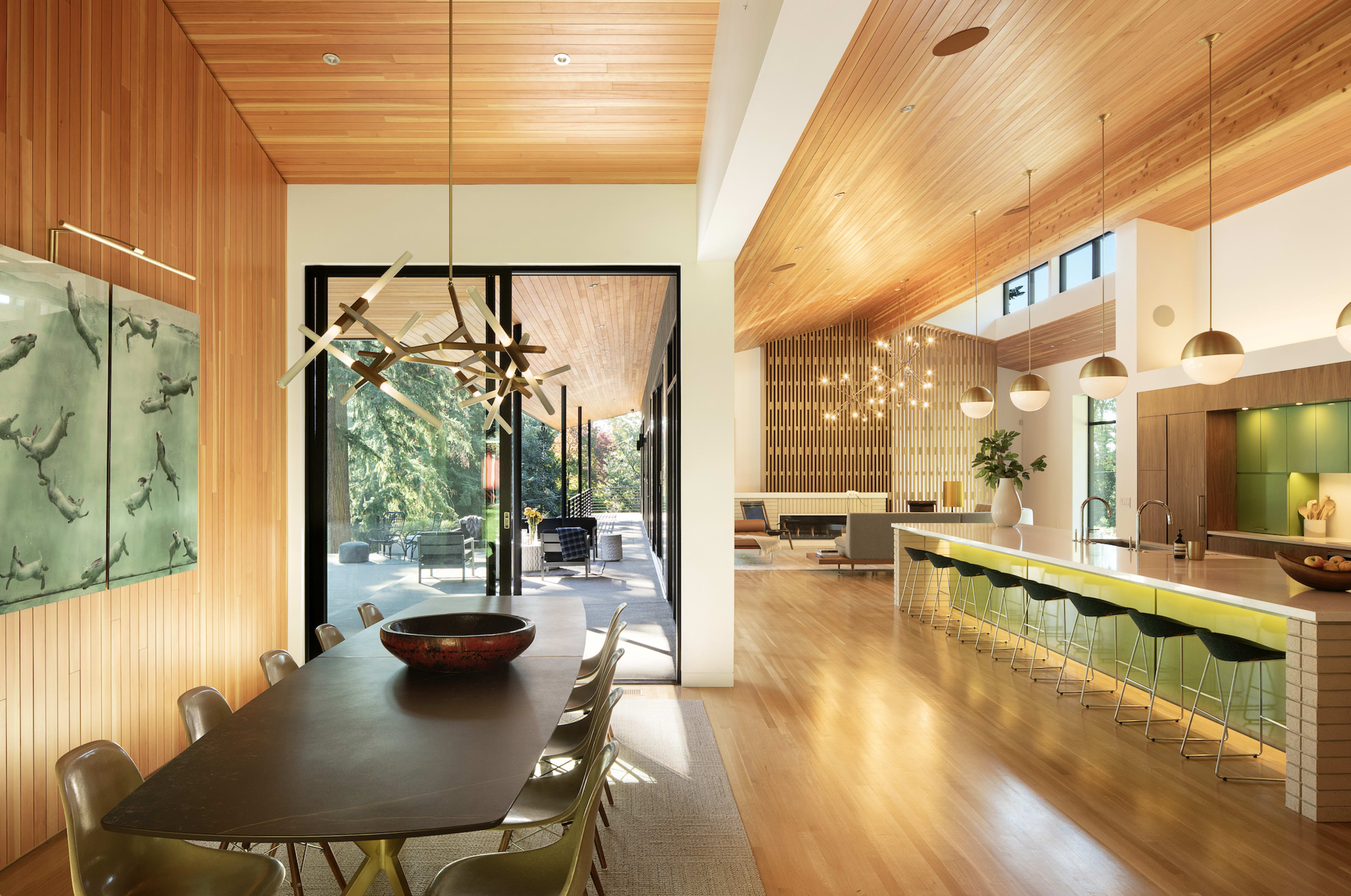 An open-concept dining area with a wooden dining table, surrounded by chairs, leading to a kitchen with bar seating and large windows overlooking a patio.
