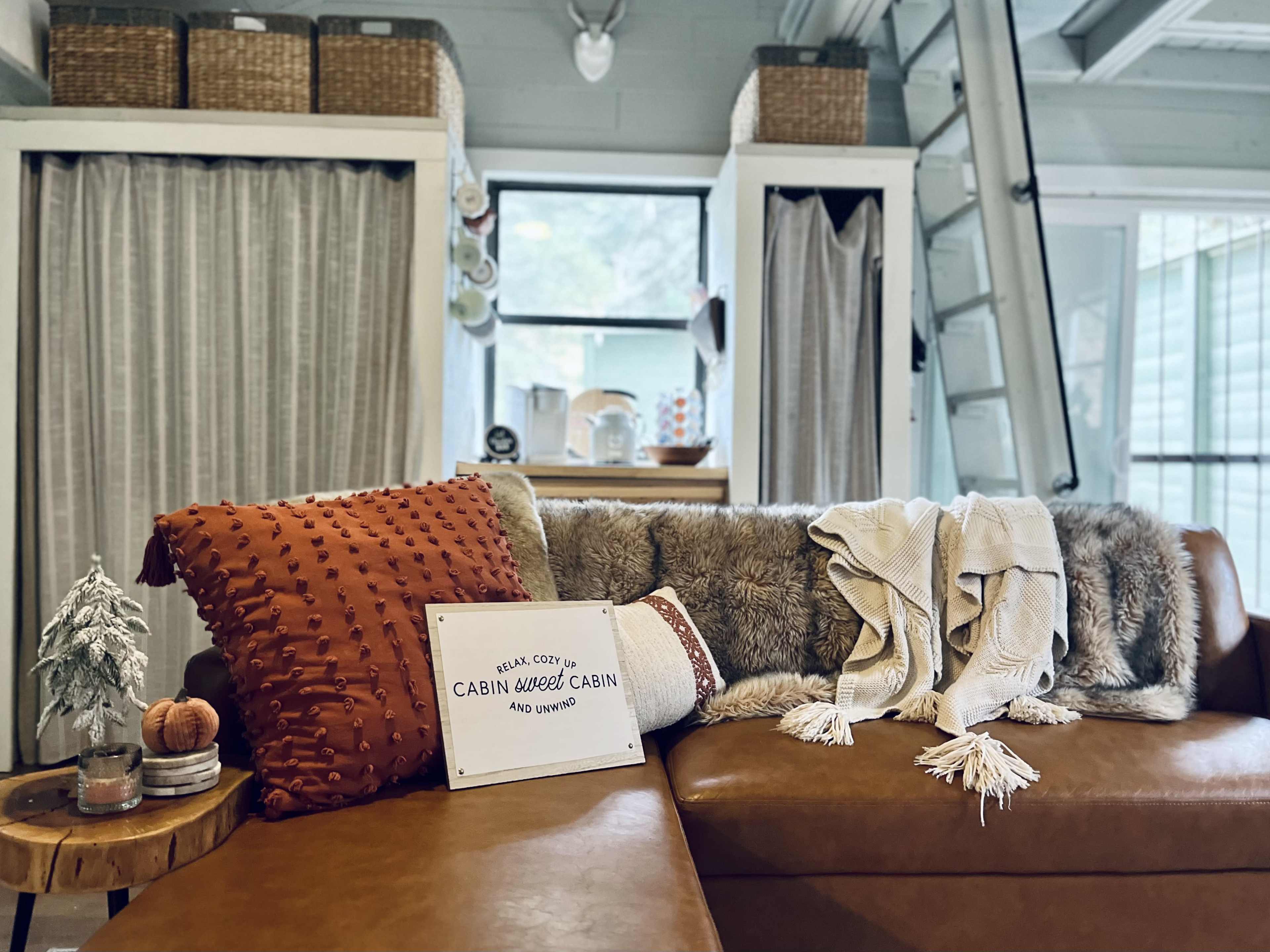 A cozy seating area features a brown leather couch adorned with decorative pillows and a blanket, alongside a sign that reads "CABIN WEED CABIN 490 W. 2ND."