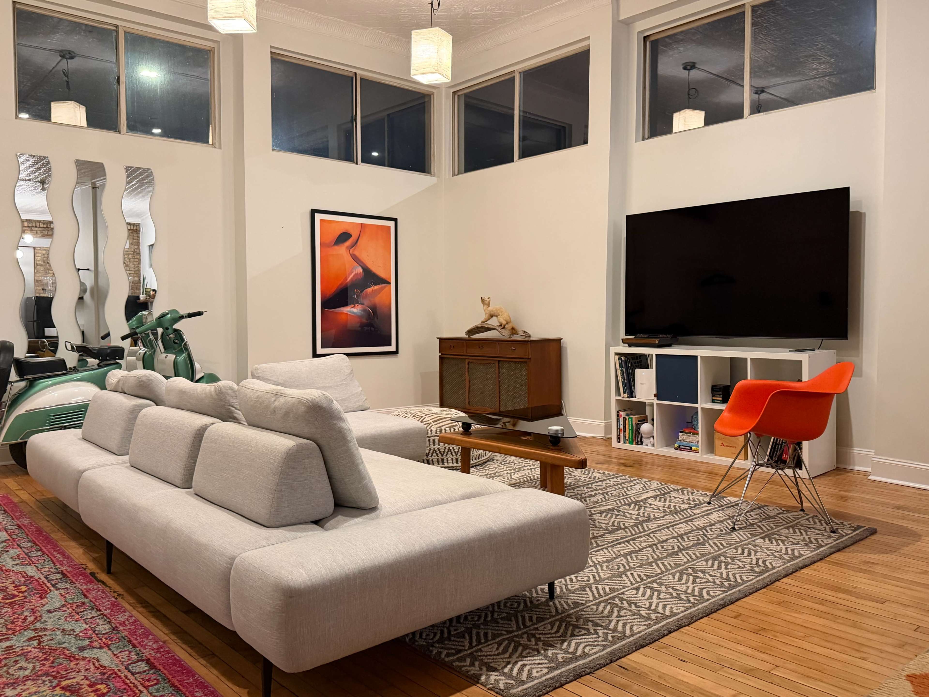 The living room features a light-colored sofa, a large television mounted on the wall, a wooden cabinet, and decorative art, with a patterned rug covering the hardwood floor.