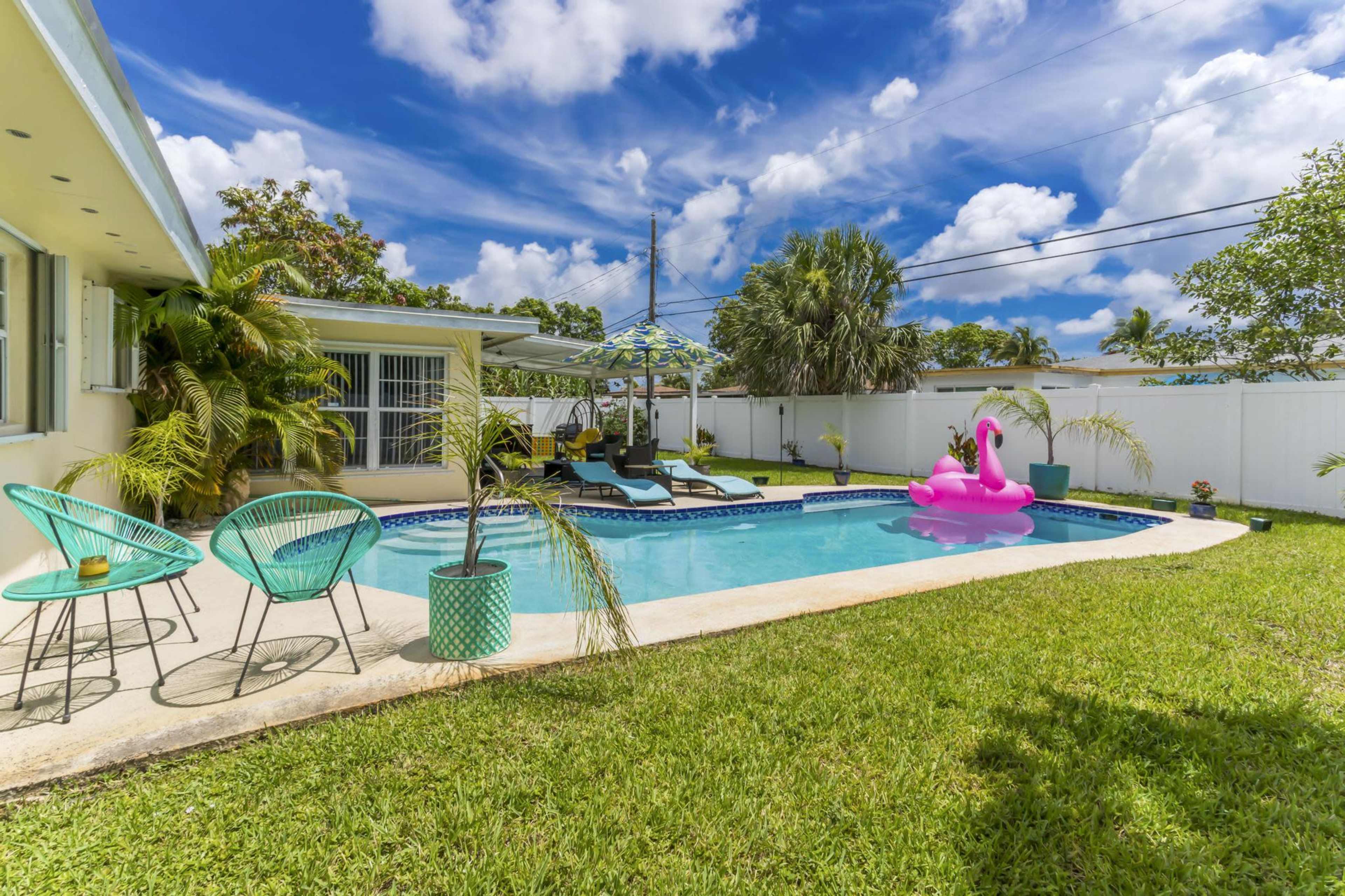 The image shows a backyard pool area featuring a swimming pool with a pink inflatable flamingo, surrounded by green grass and patio furniture.