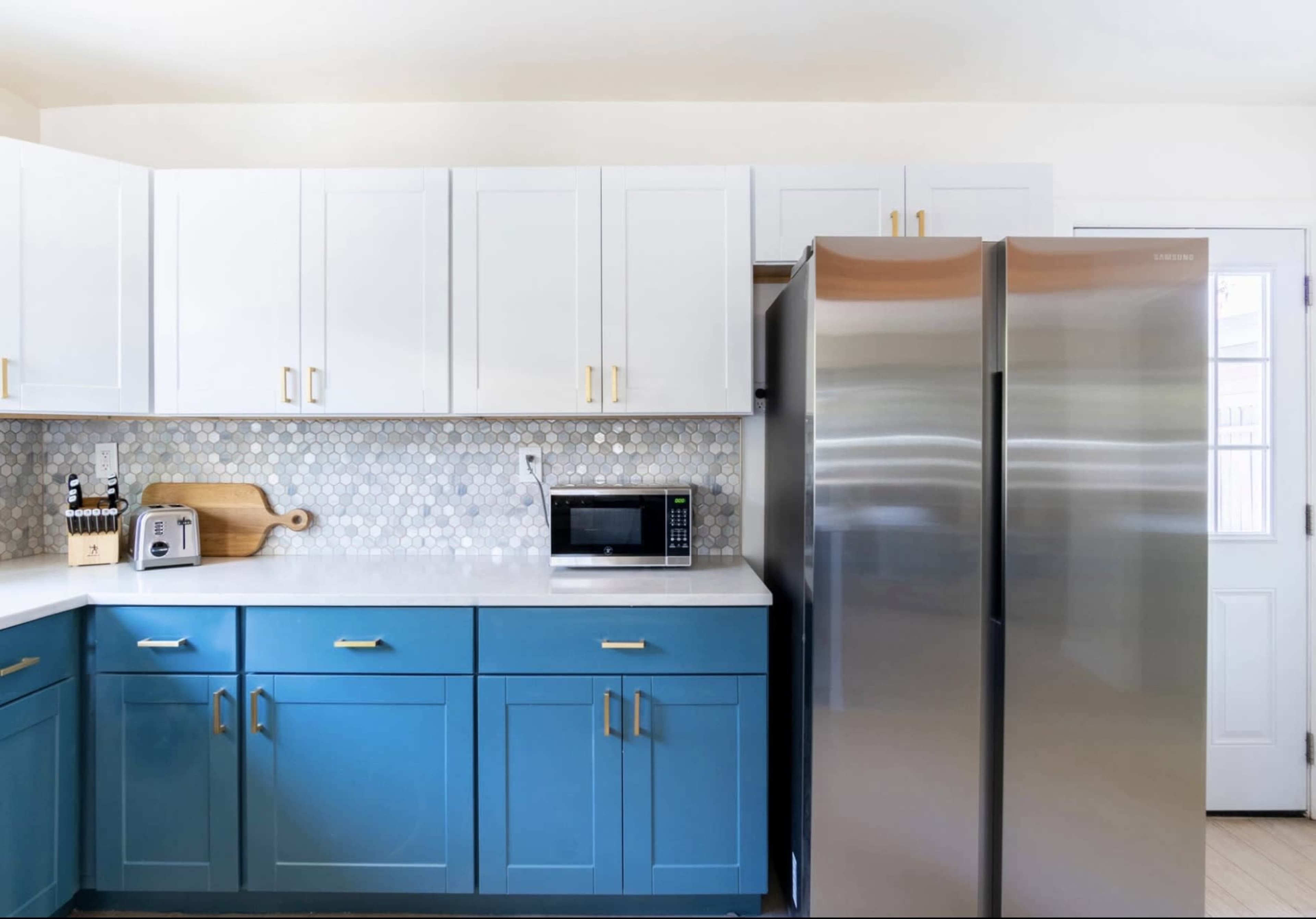 A modern kitchen with blue and white cabinets, a stainless steel refrigerator, a microwave, and a hexagon tile backsplash.