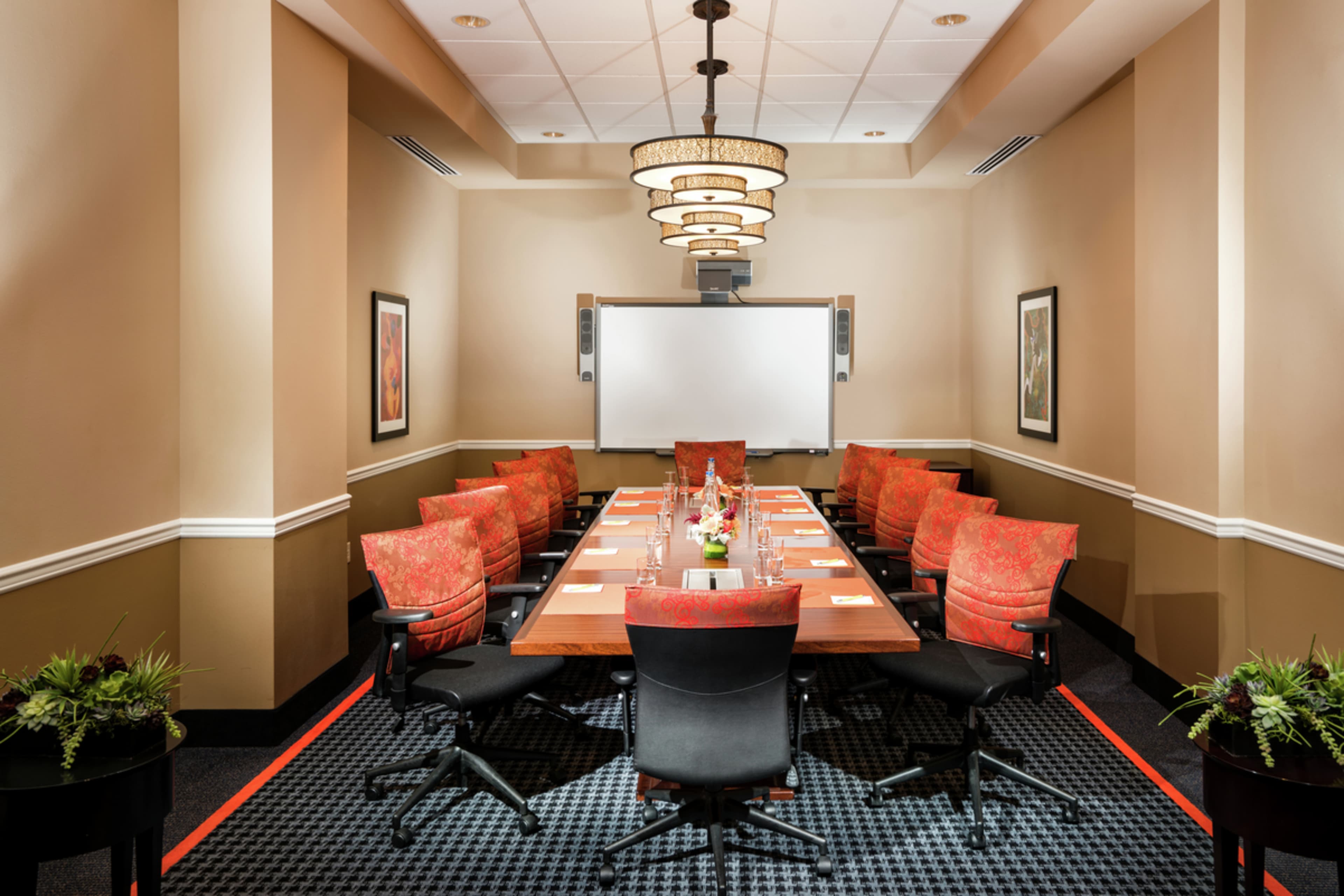 A conference room features a large rectangular table surrounded by upholstered chairs and a presentation screen at the front.