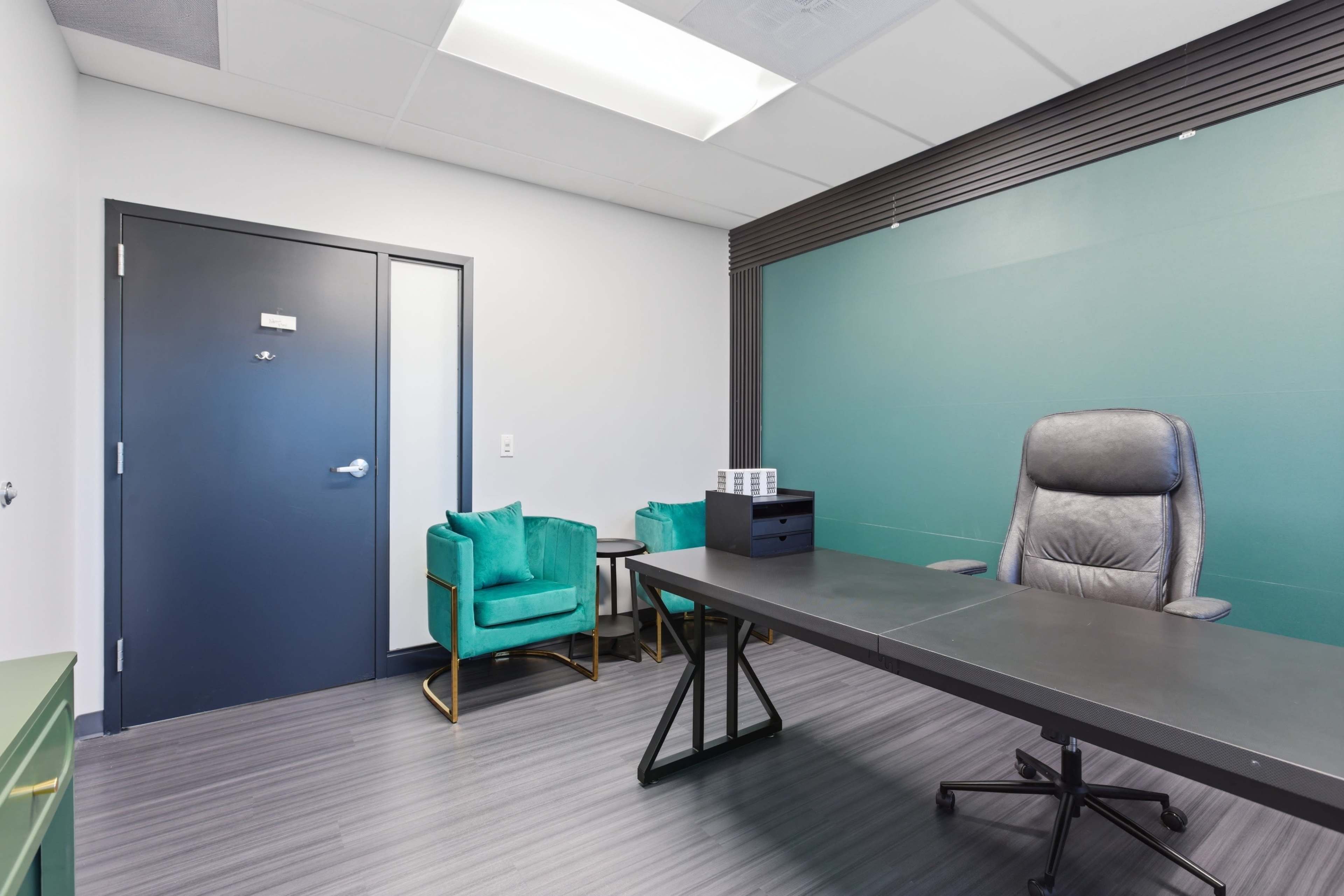 The image shows a modern office space featuring a large desk, gray ergonomic chair, green accent wall, and two teal chairs beside a small table.