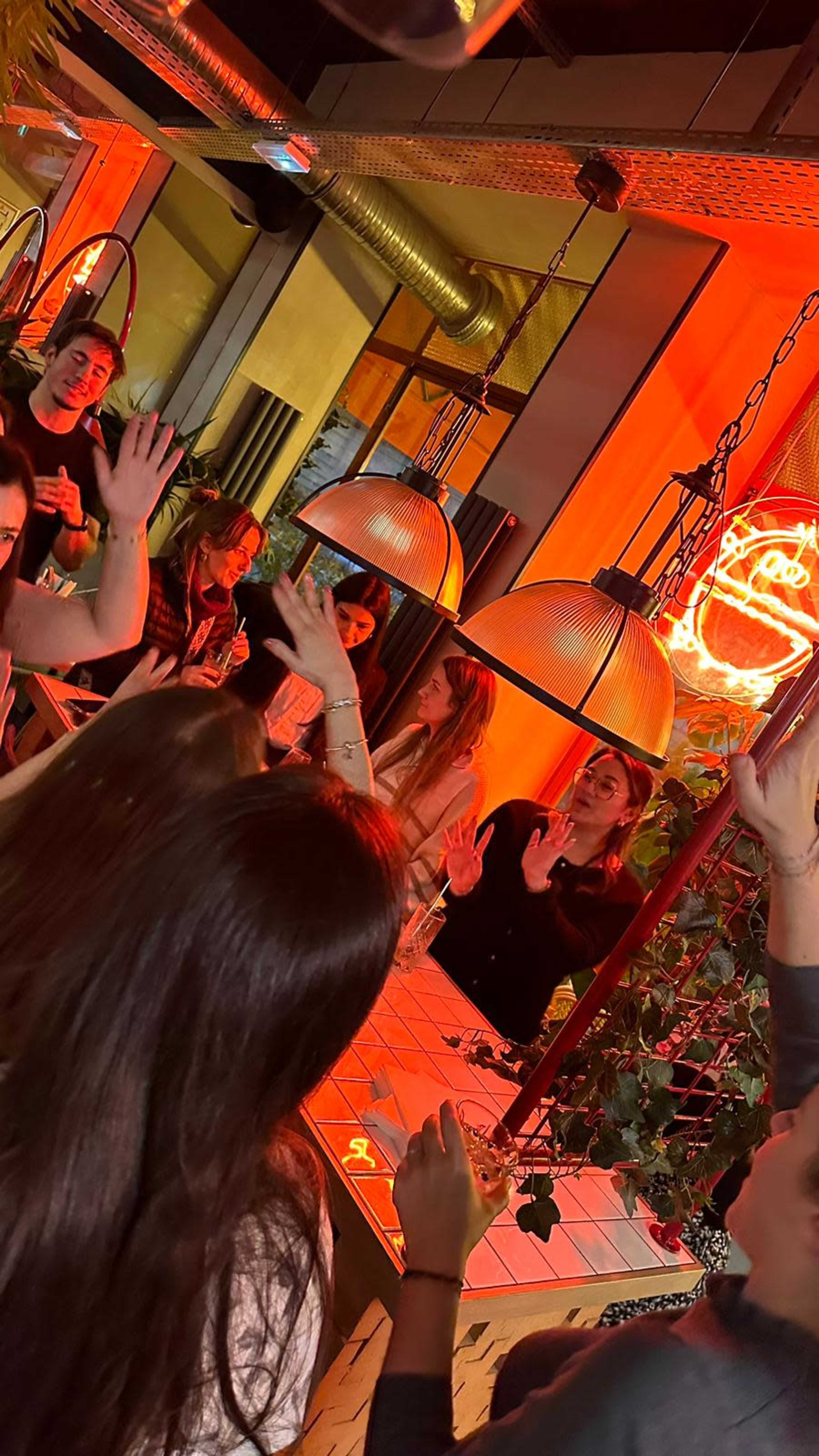 A group of people raises their hands and interacts enthusiastically in a lively, illuminated setting with hanging lights and a neon sign.